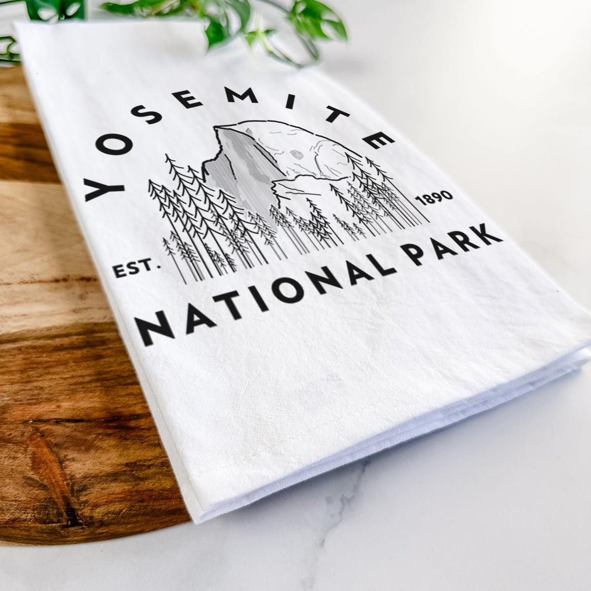 Close-up of a folded white tea towel on a wooden board, showing a round badge of Half Dome and pine trees, with the words Yosemite National Park, Est. 1890.