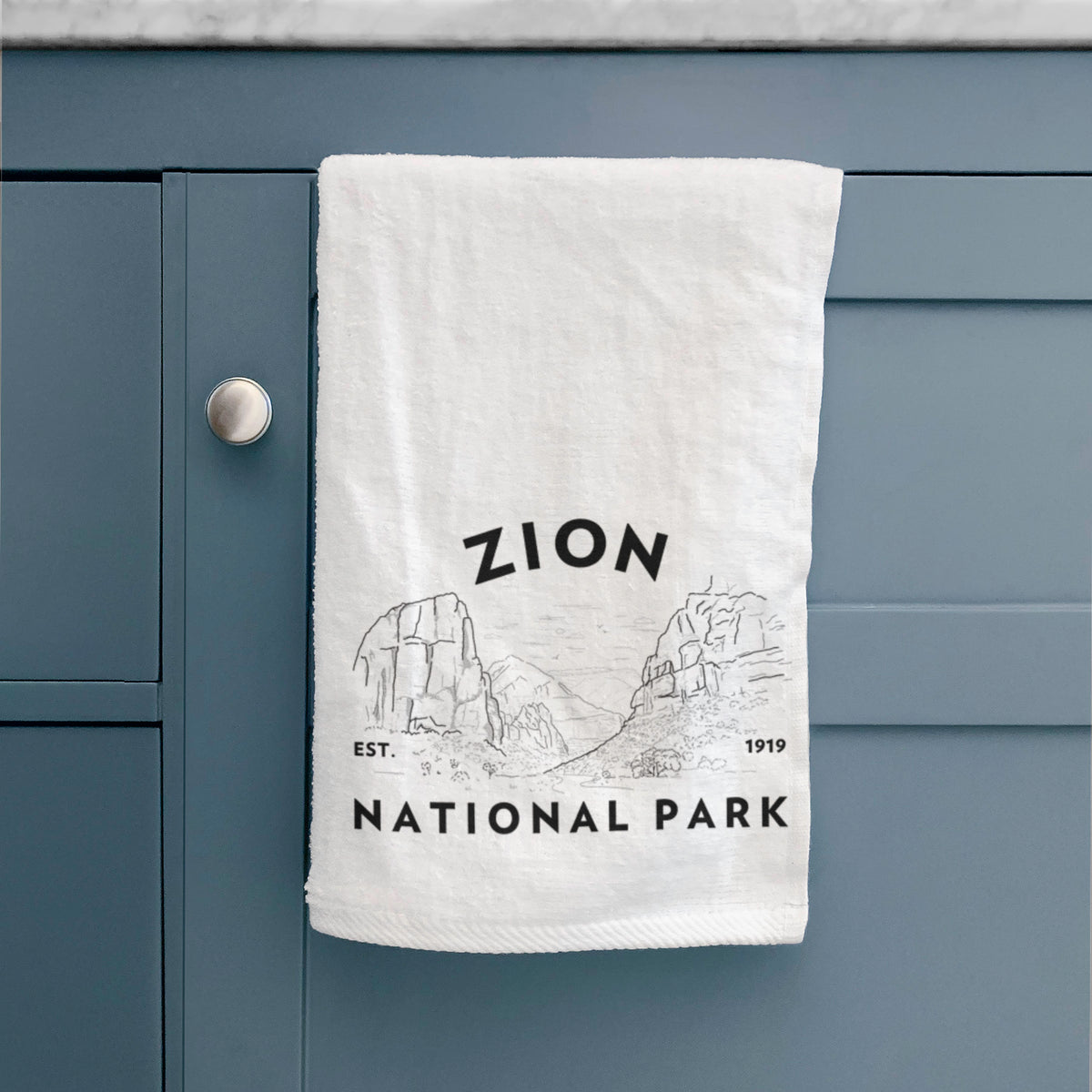 White hand towel printed with a round badge of canyon walls, with the words Zion National Park, Est. 1919, hanging on a cabinet door.