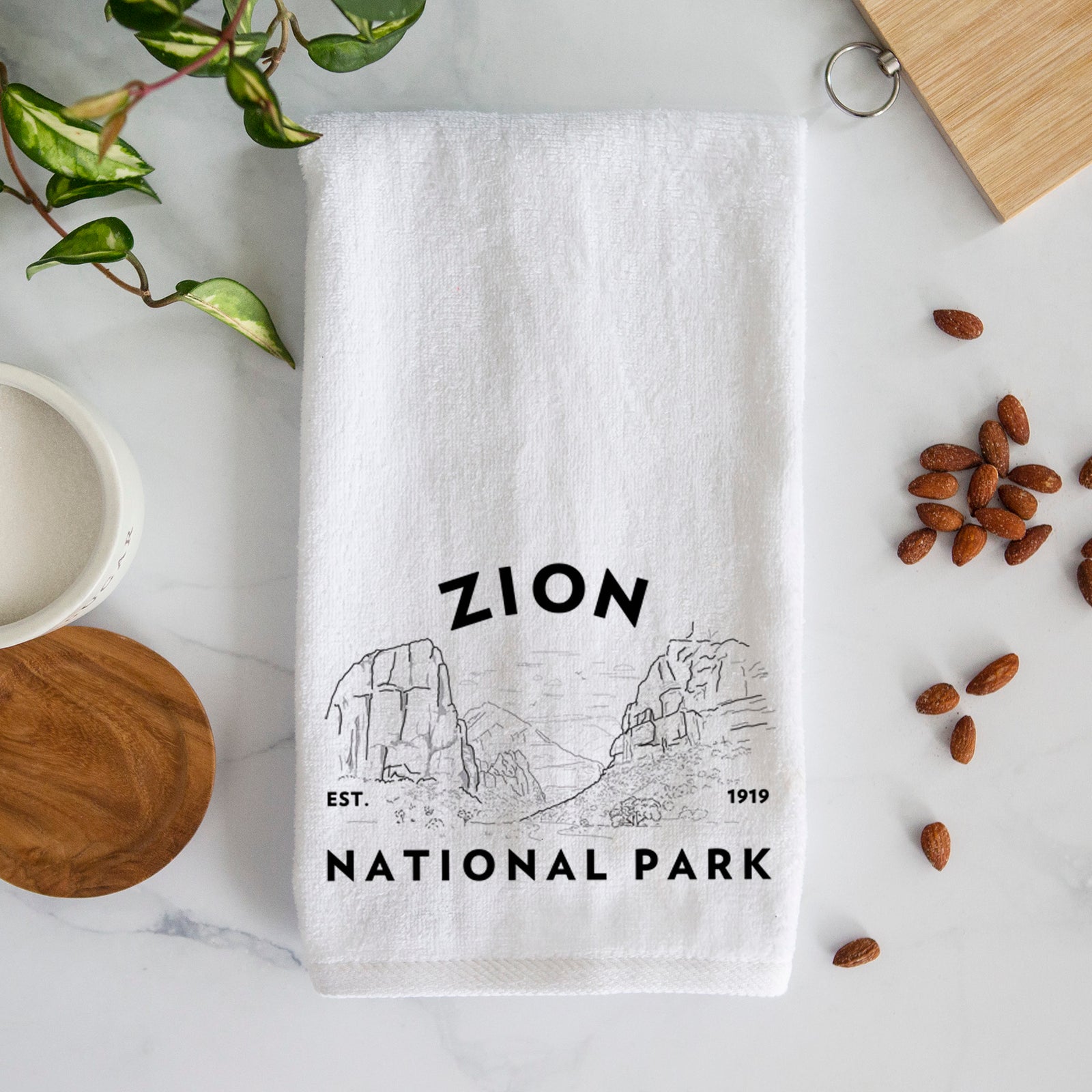 White hand towel printed with a round badge of canyon walls, with the words Zion National Park, Est. 1919.