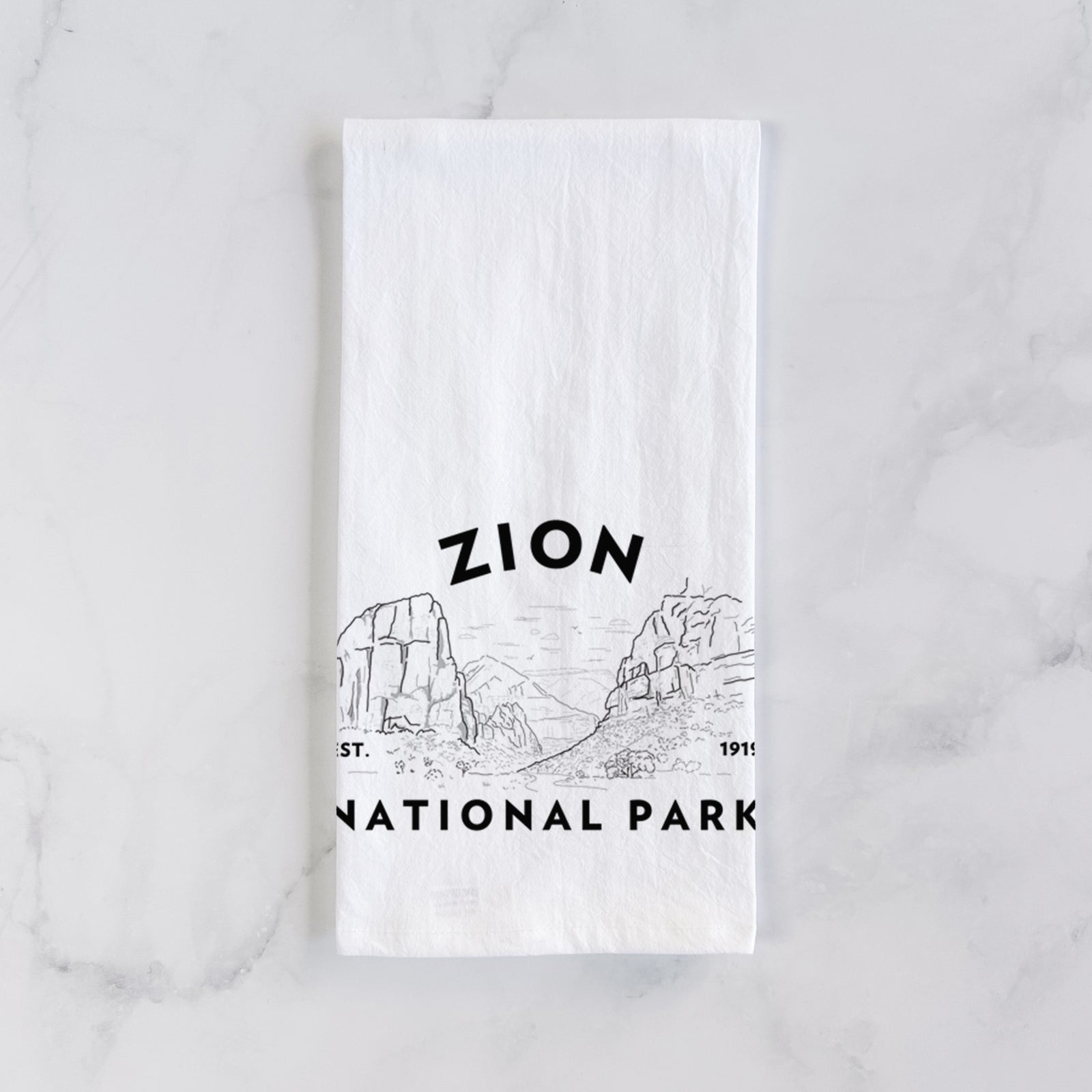 White cotton tea towel printed with a round badge of canyon walls, with the words Zion National Park, Est. 1919, on a marble counter beside a round wooden cutting board and greenery.
