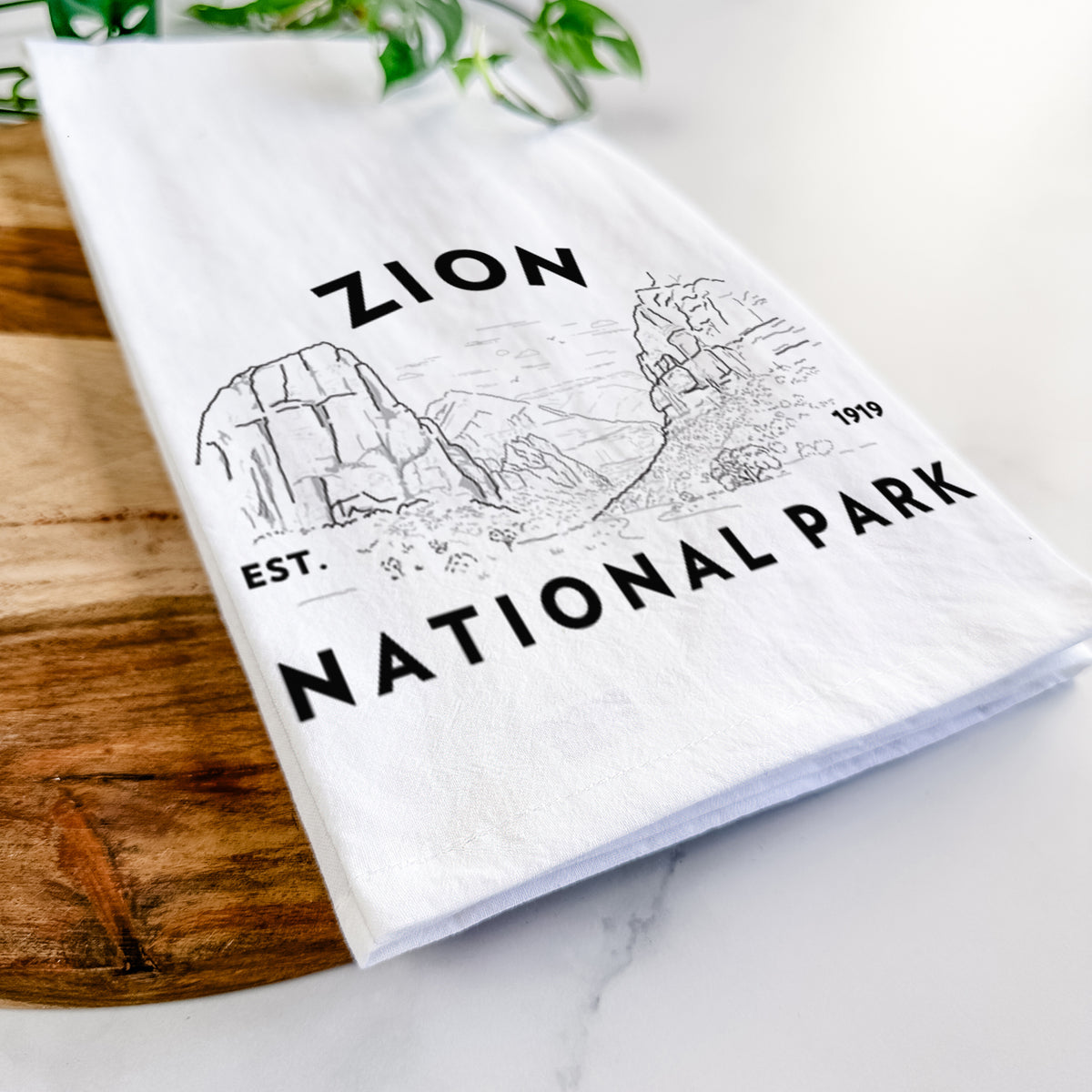 Close-up of a folded white tea towel on a wooden board, showing a round badge of canyon walls, with the words Zion National Park, Est. 1919.