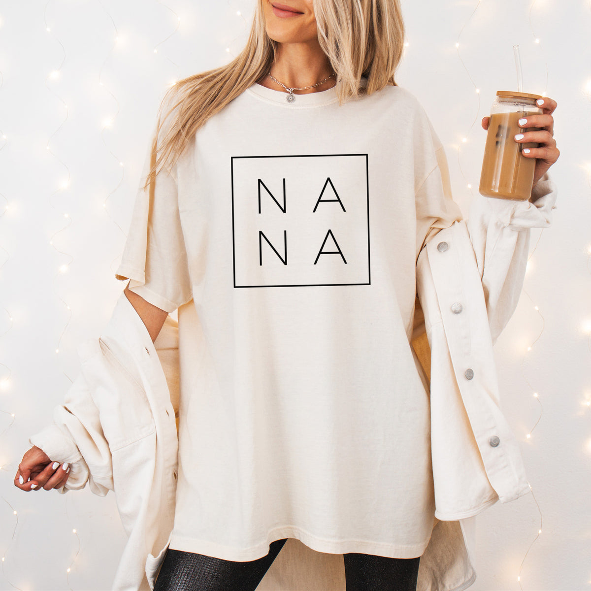 Woman wearing a Comfort Colors t-shirt printed with the word NANA in thin black letters stacked inside a square outline, outdoors.