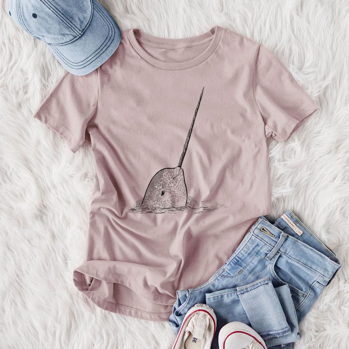 Women's relaxed fit cotton t-shirt printed with a black ink illustration of a narwhal with its long spiral tusk pointing upward, styled flat on a white faux fur rug with jeans.