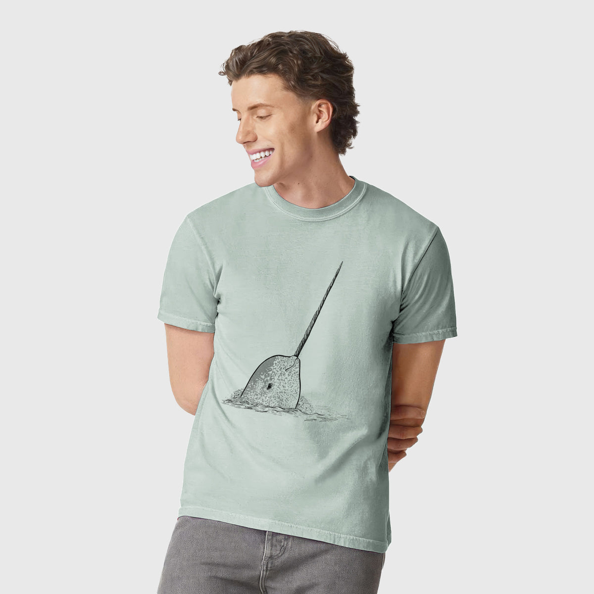 Man wearing a Comfort Colors t-shirt printed with a black ink illustration of a narwhal with its long spiral tusk pointing upward.