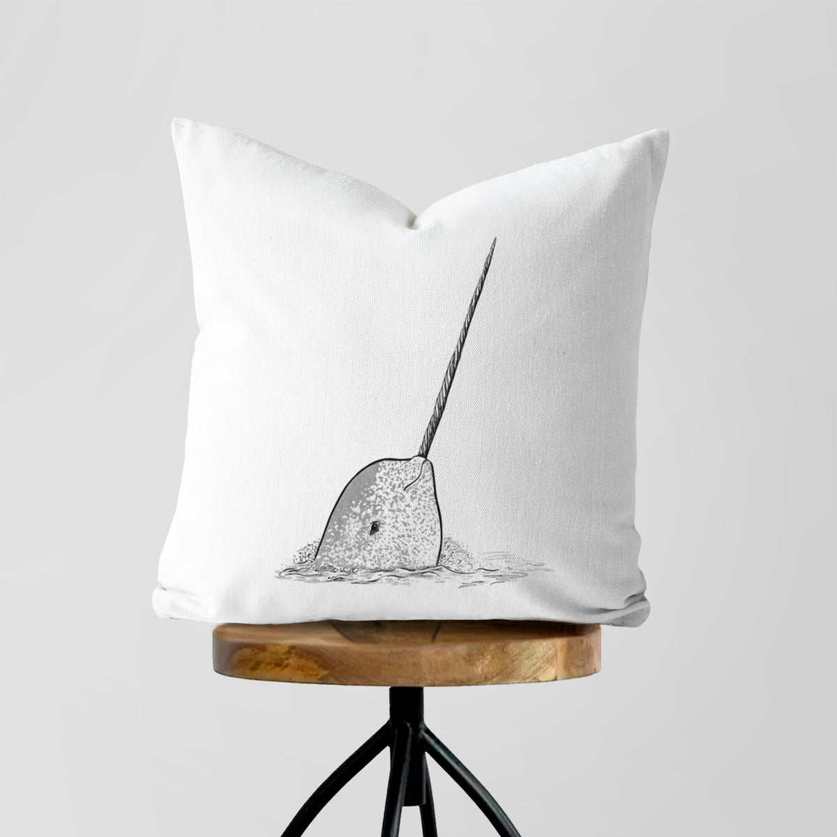 White pillow cover printed with a black ink illustration of a narwhal with its long spiral tusk pointing upward, on a wooden stool.