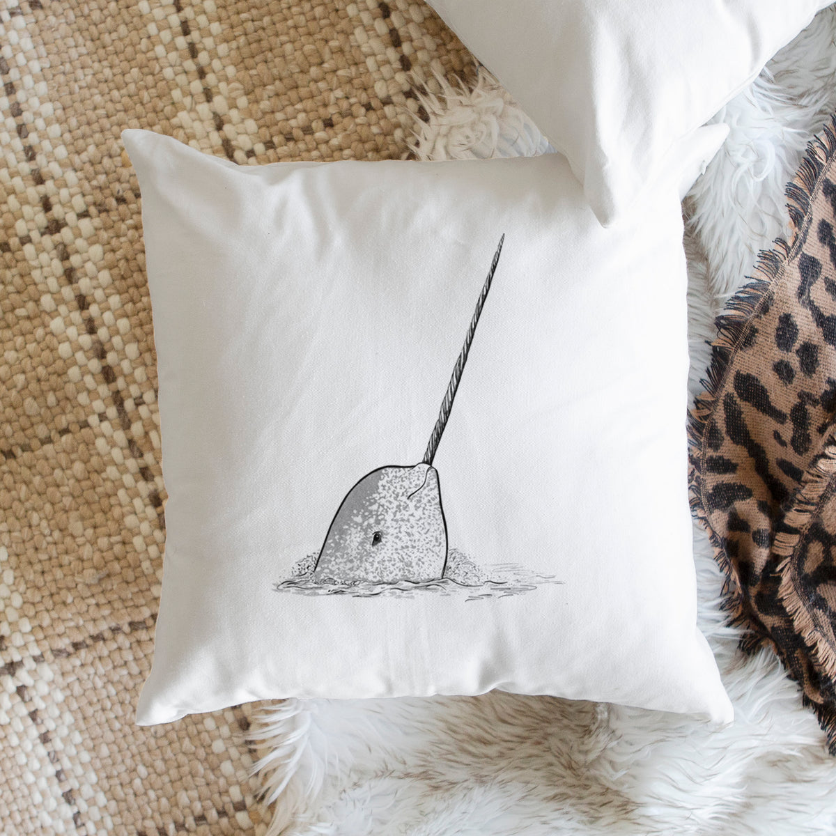 White pillow cover printed with a black ink illustration of a narwhal with its long spiral tusk pointing upward, on a woven chair.