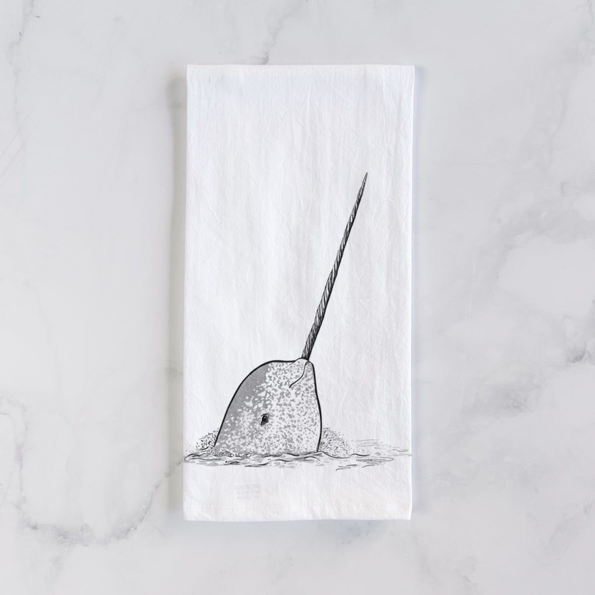 White cotton tea towel laid flat on marble, printed with a black ink illustration of a narwhal with its long spiral tusk pointing upward.