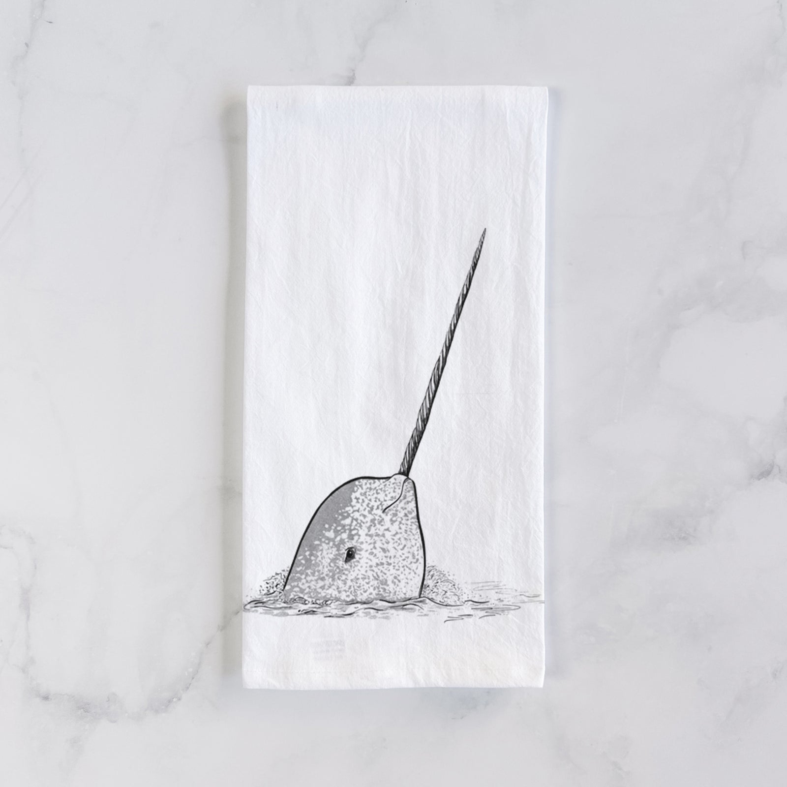 White cotton tea towel printed with a black ink illustration of a narwhal with its long spiral tusk pointing upward, on a marble counter beside a round wooden cutting board and greenery.