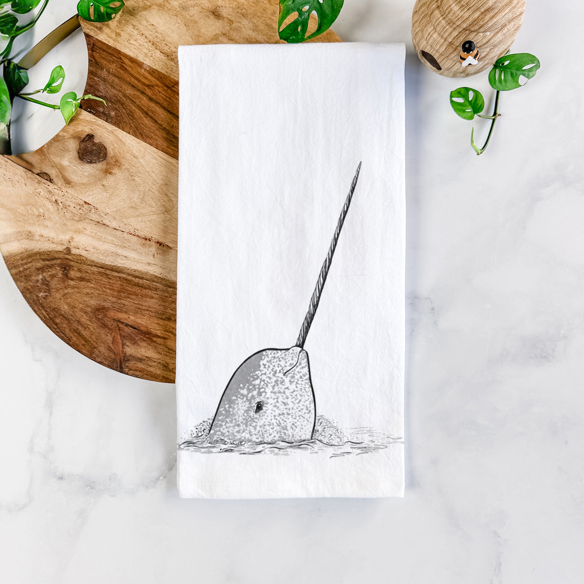 White cotton tea towel printed with a black ink illustration of a narwhal with its long spiral tusk pointing upward, on a marble counter beside a round wooden cutting board and greenery.