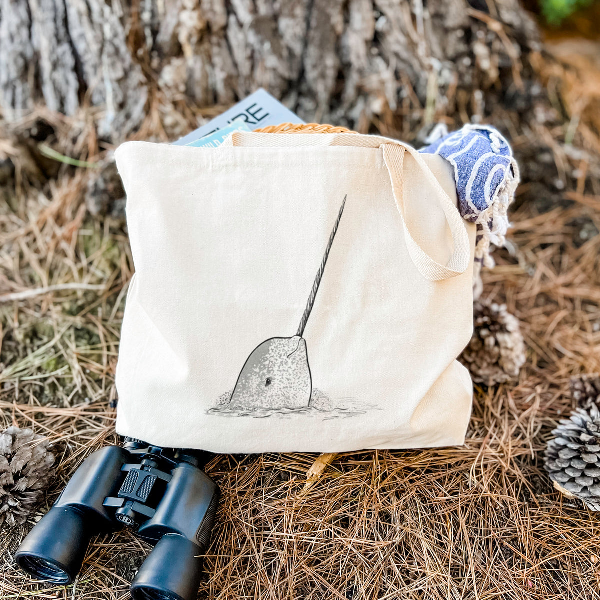 Canvas tote bag printed with a black ink illustration of a narwhal with its long spiral tusk pointing upward, resting on dry grass beside a pair of binoculars.