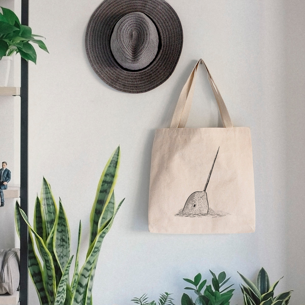 Canvas tote bag printed with a black ink illustration of a narwhal with its long spiral tusk pointing upward, hanging on a wall hook below a hat, next to potted plants.