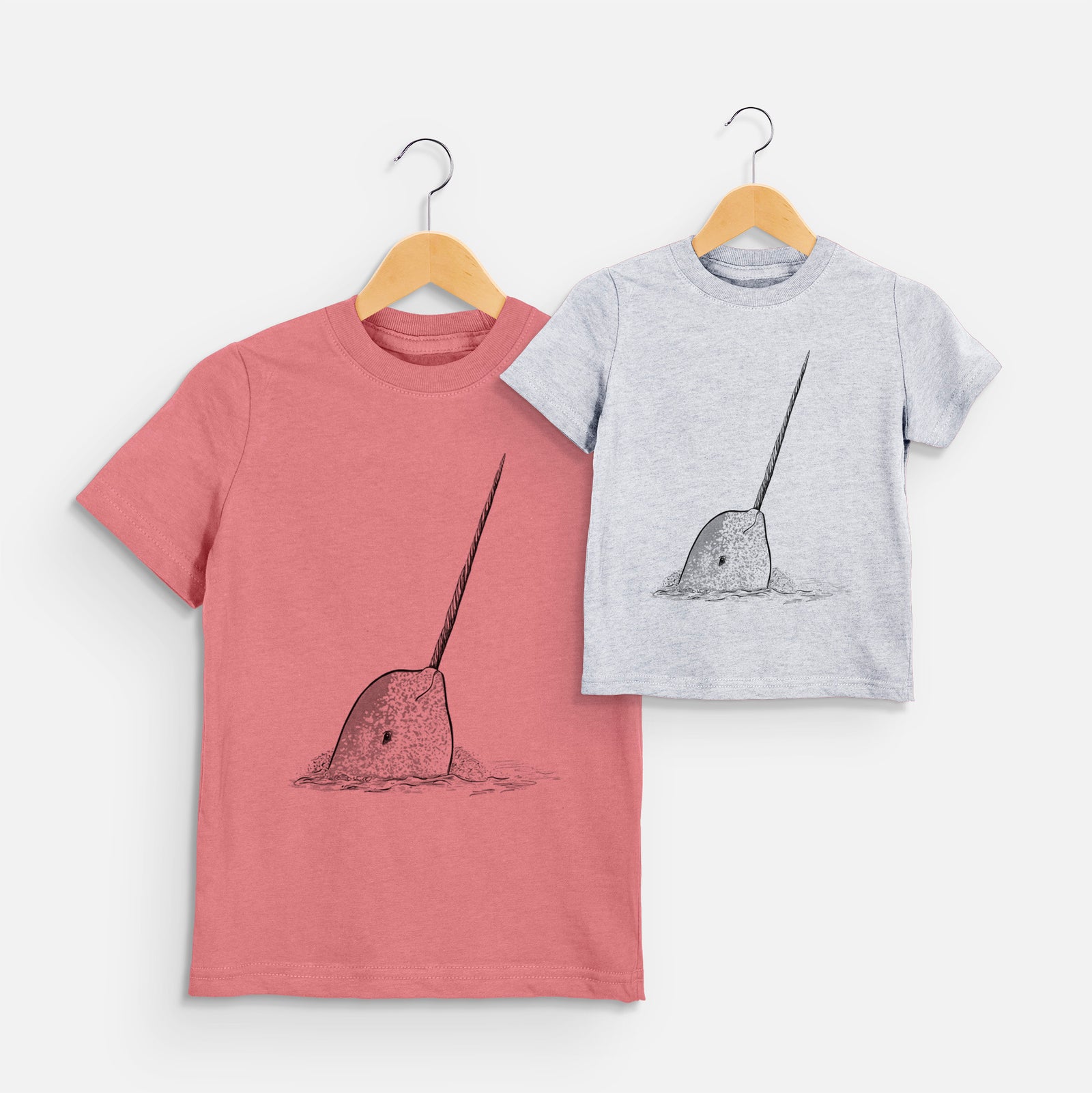 Two youth t-shirts in different colors on hangers, printed with a black ink illustration of a narwhal with its long spiral tusk pointing upward.