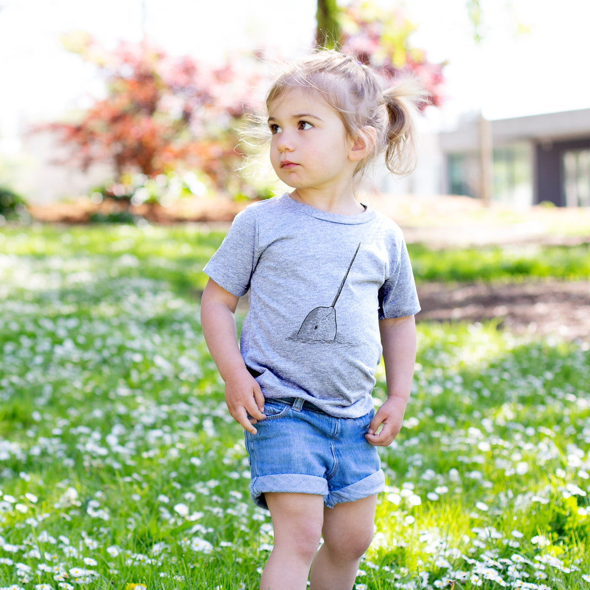 Toddler girl walking outdoors wearing a t-shirt printed with a black ink illustration of a narwhal with its long spiral tusk pointing upward.