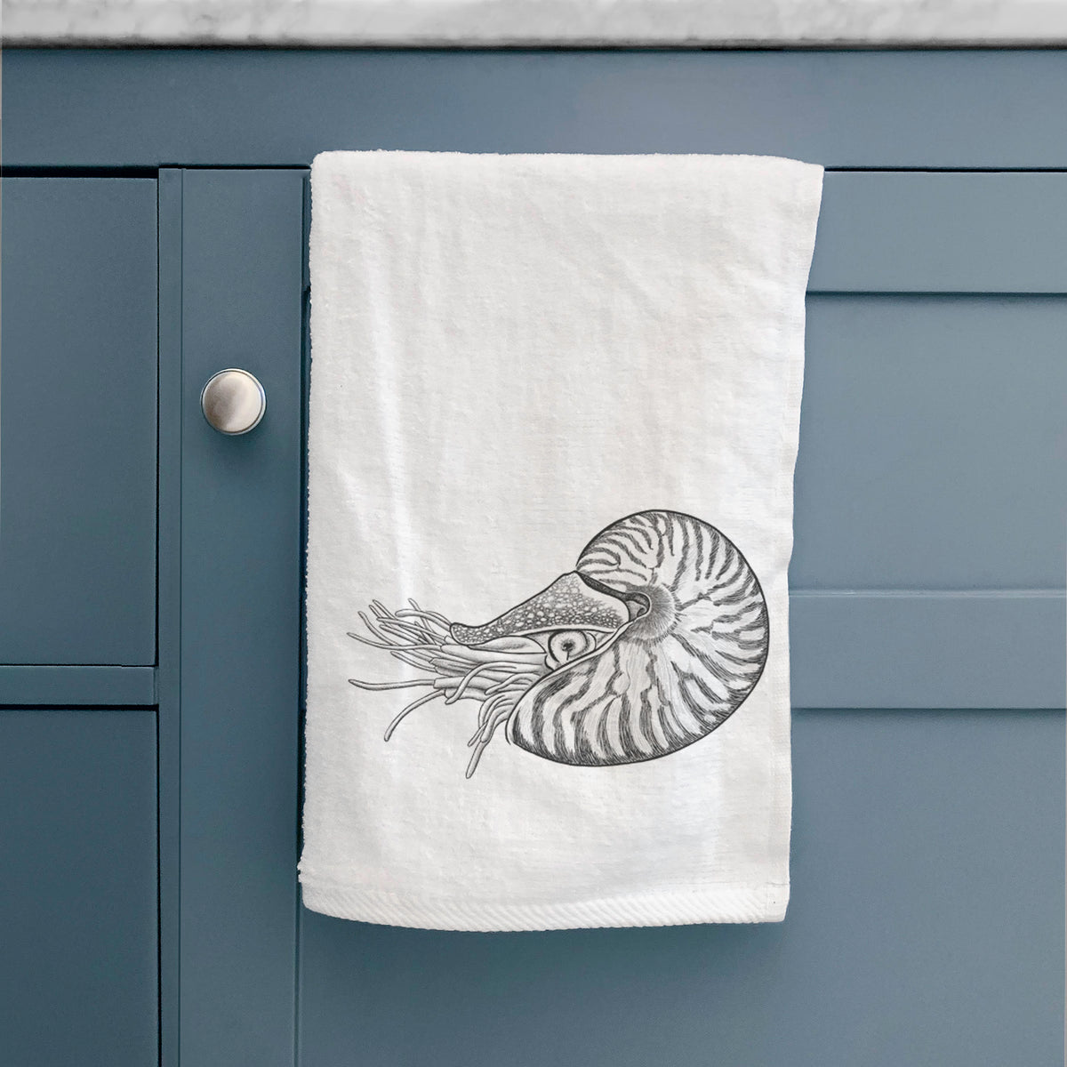 White hand towel printed with a detailed black ink illustration of a chambered nautilus with its striped spiral shell and tentacles, hanging on a cabinet door.