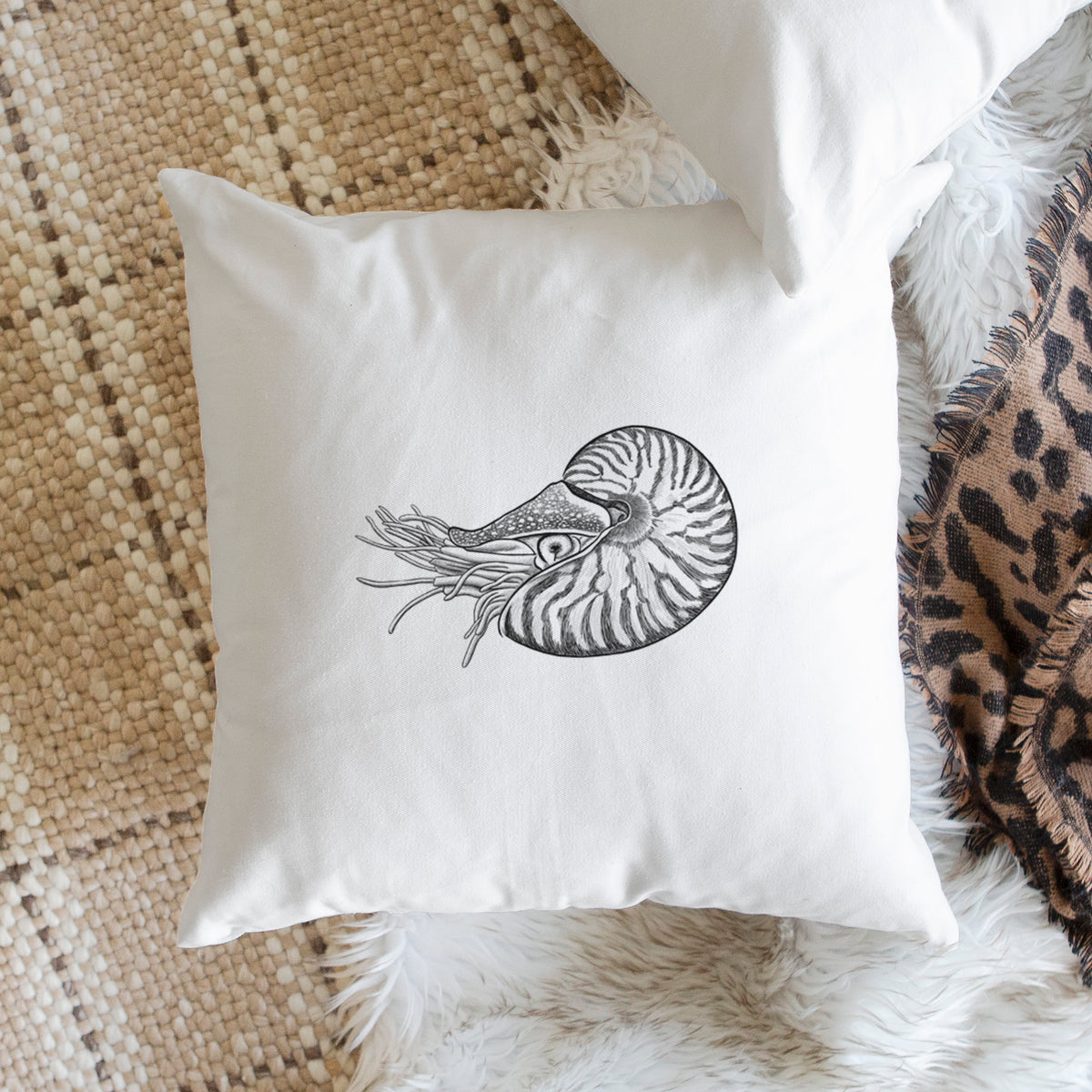 White pillow cover printed with a detailed black ink illustration of a chambered nautilus with its striped spiral shell and tentacles, on a woven chair.