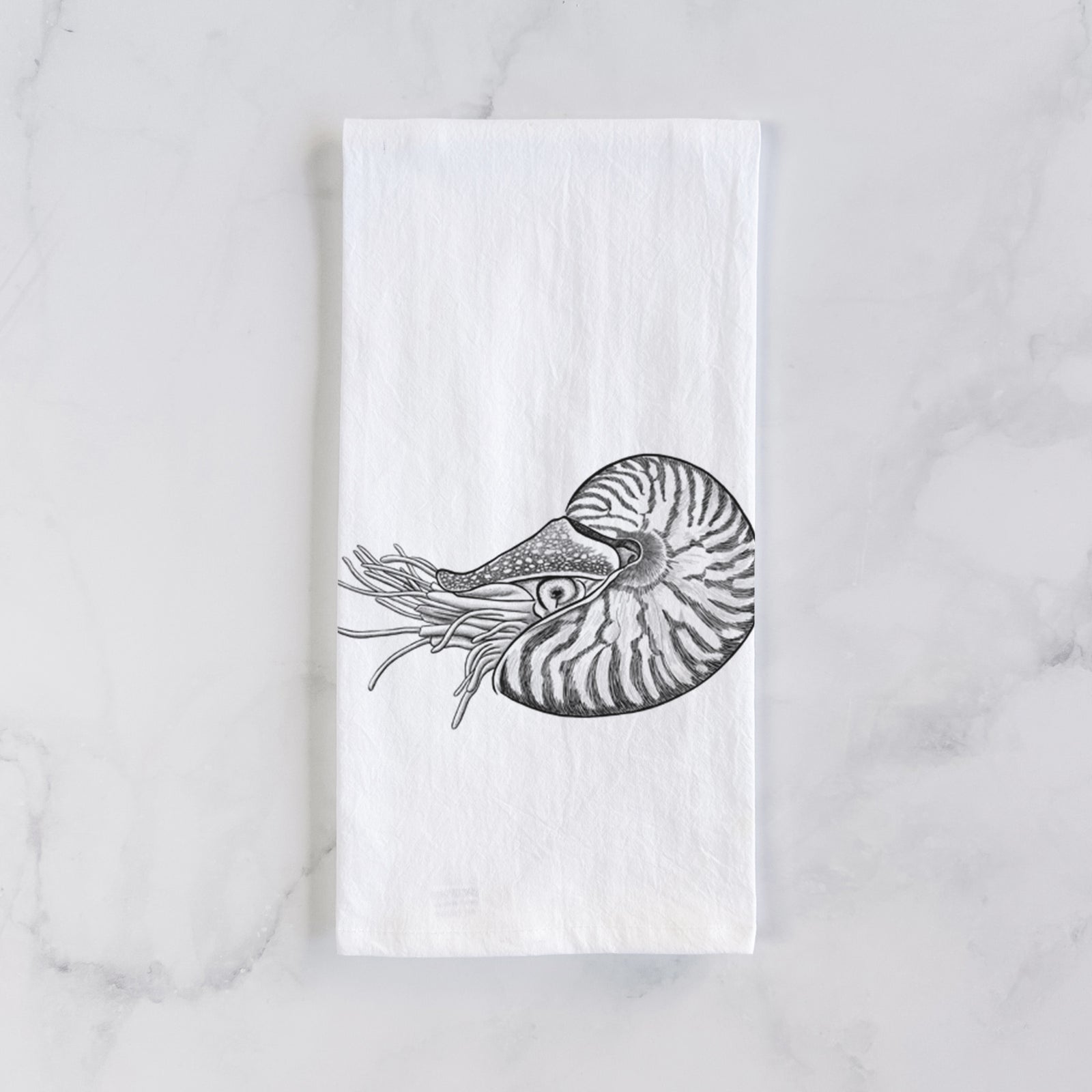 White cotton tea towel printed with a detailed black ink illustration of a chambered nautilus with its striped spiral shell and tentacles, on a marble counter beside a round wooden cutting board and greenery.