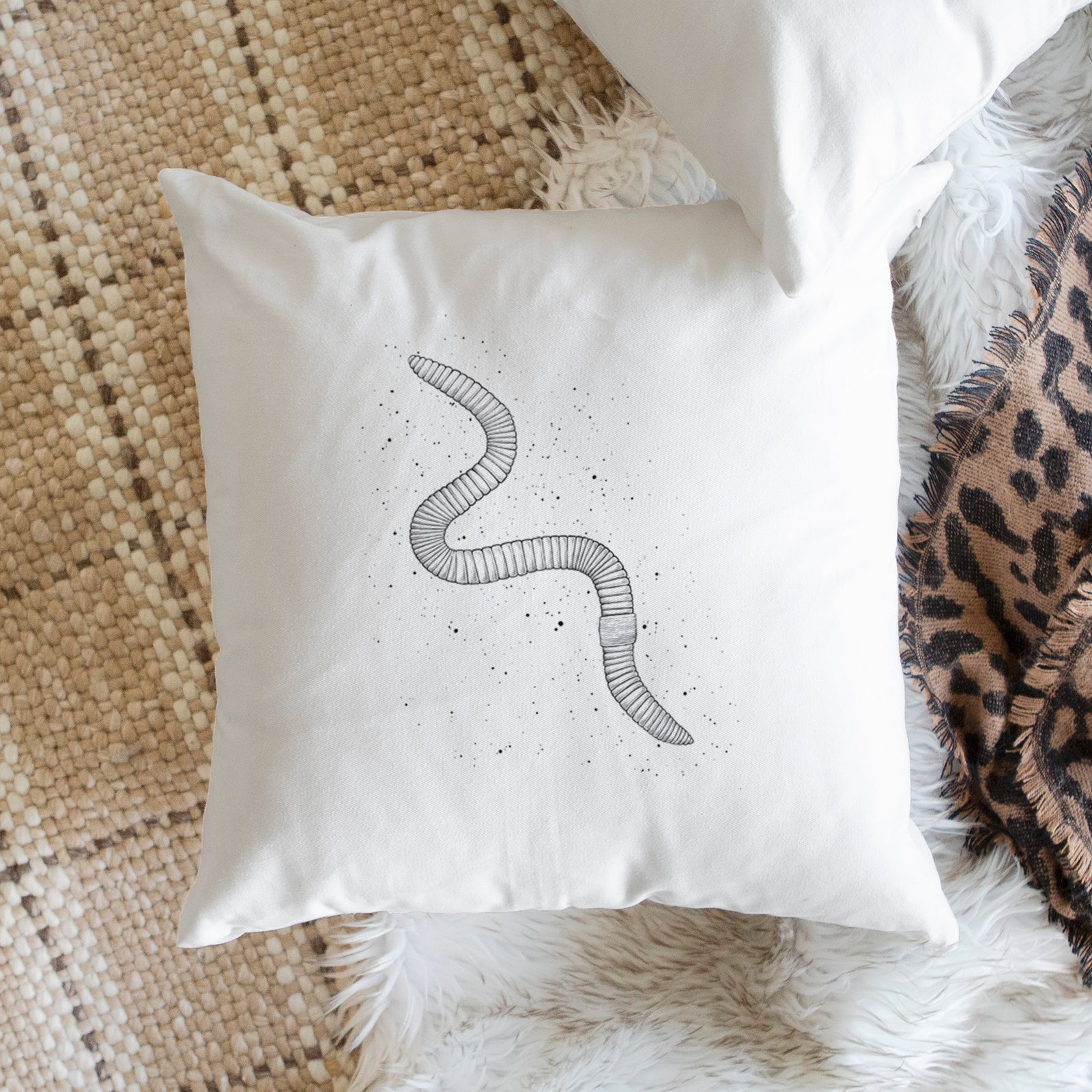 White pillow cover printed with a black ink illustration of a nightcrawler earthworm with soil specks, on a bench beside plants.