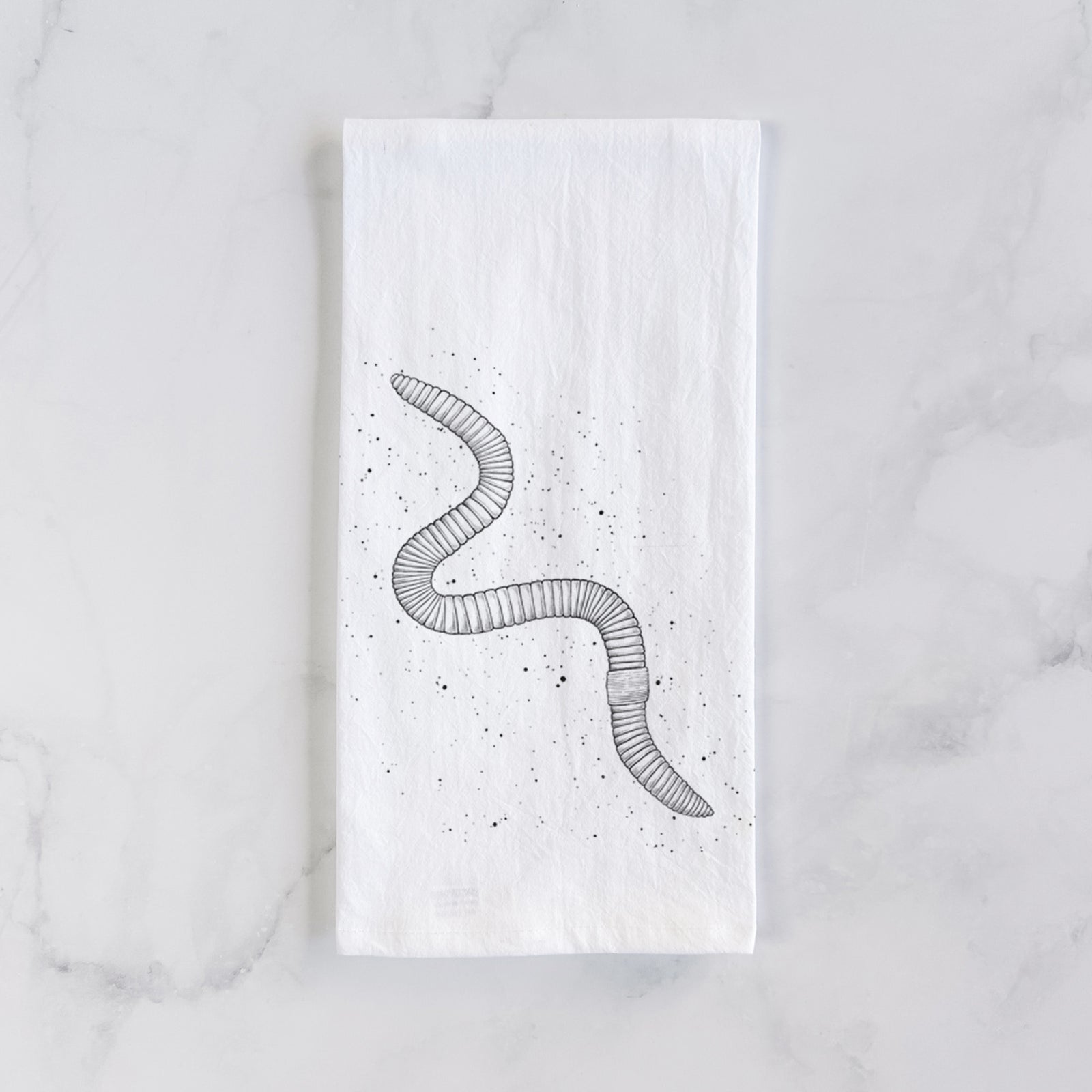 White cotton tea towel printed with a black ink illustration of a nightcrawler earthworm with soil specks, on a marble counter beside a round wooden cutting board and greenery.