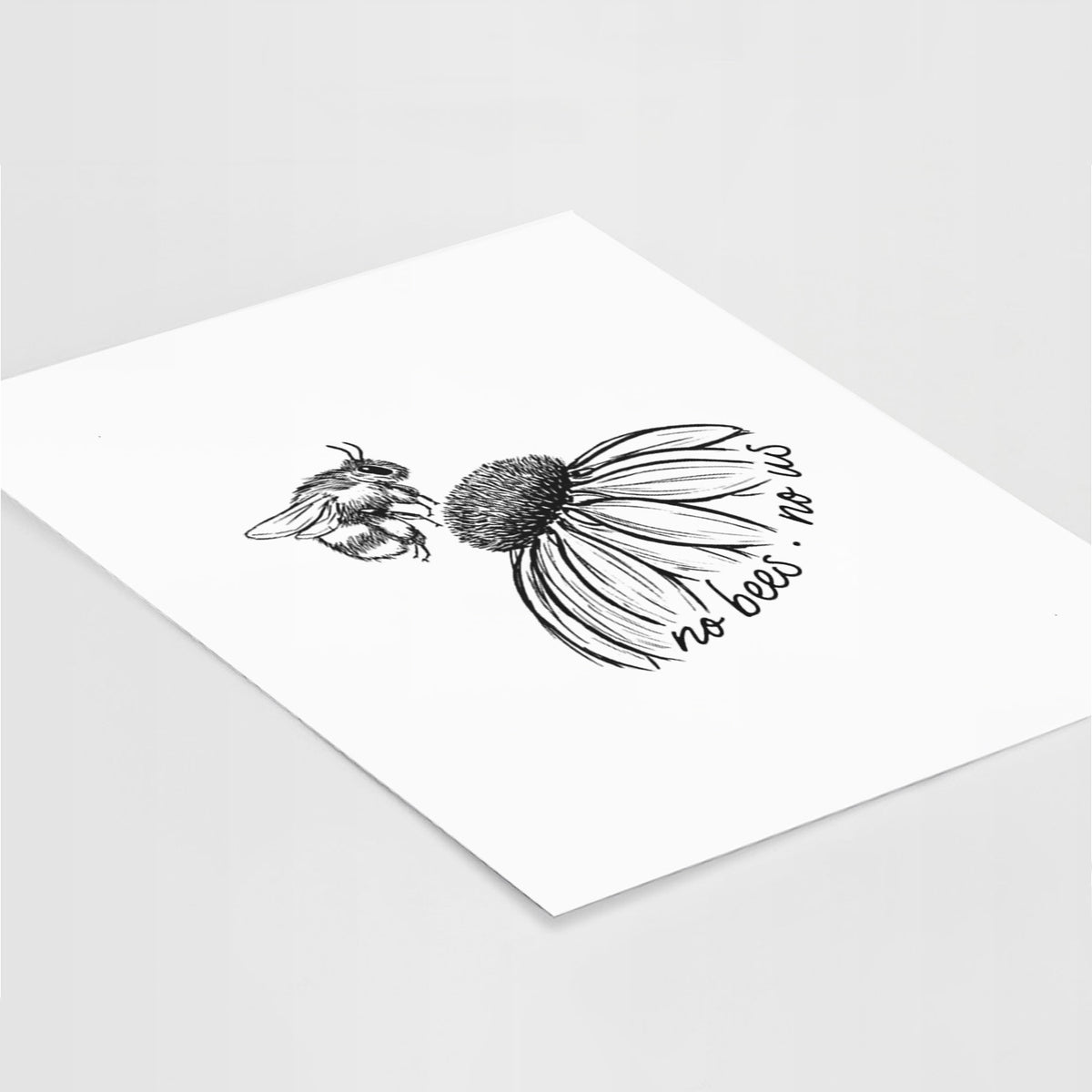 Art print of a black ink illustration of a bee flying toward a coneflower, with the handwritten words no bees no us, lying at an angle on a white surface.