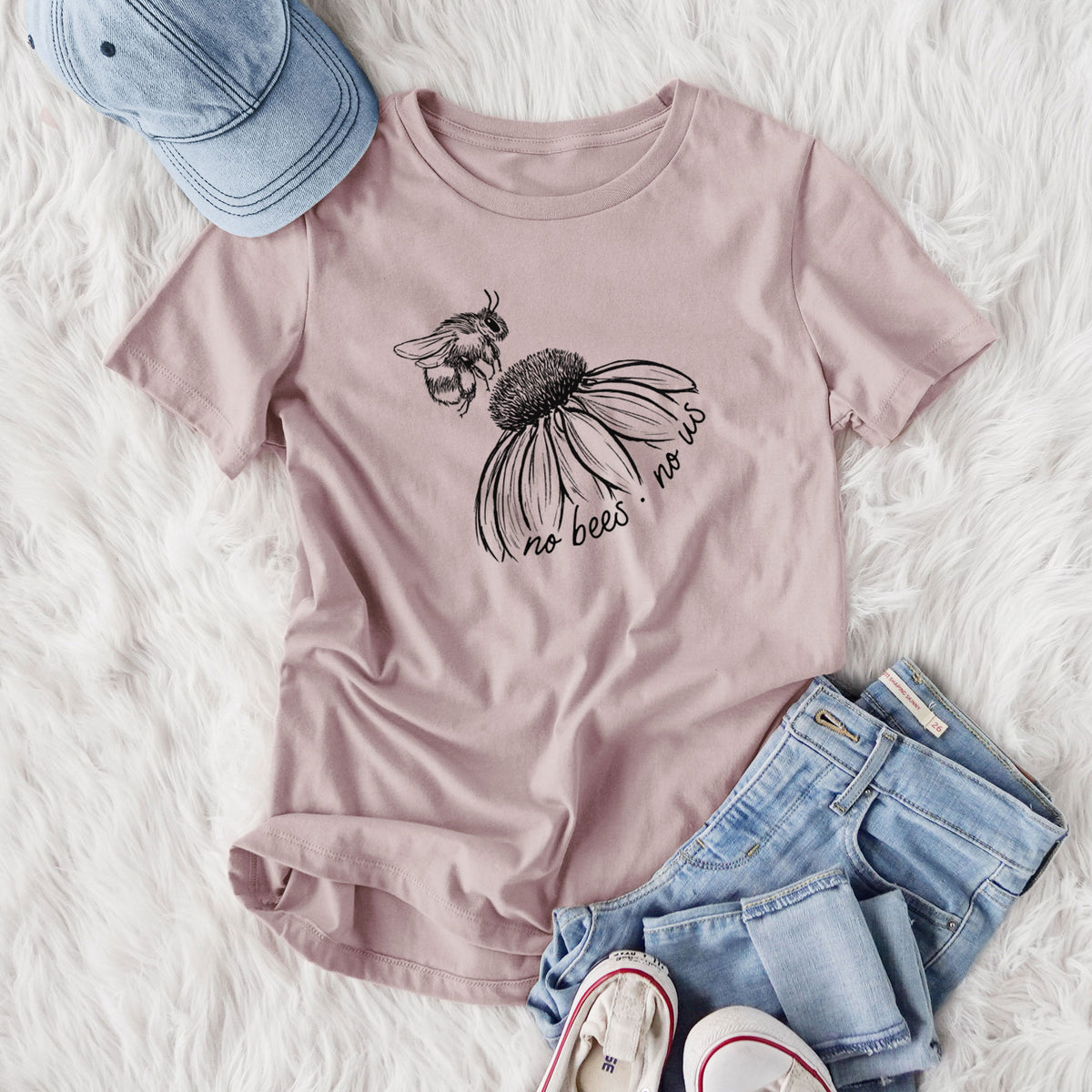 Women's relaxed fit cotton t-shirt printed with a black ink illustration of a bee flying toward a coneflower, with the handwritten words no bees no us, styled flat on a white faux fur rug with jeans.