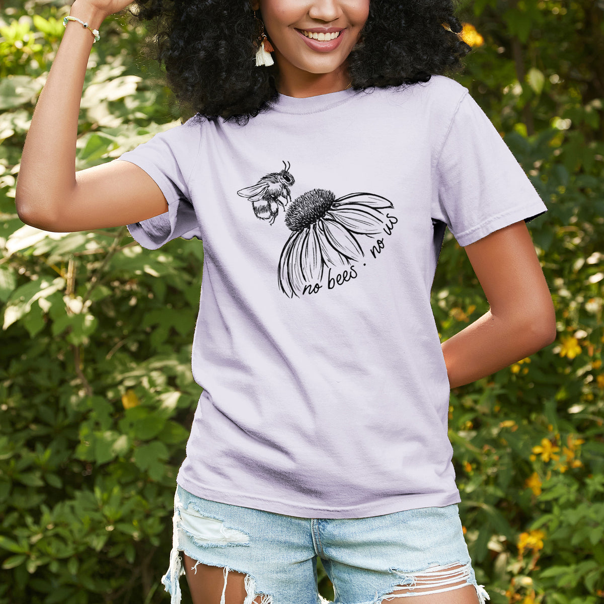 Woman wearing a Comfort Colors t-shirt printed with a black ink illustration of a bee flying toward a coneflower, with the handwritten words no bees no us, outdoors.