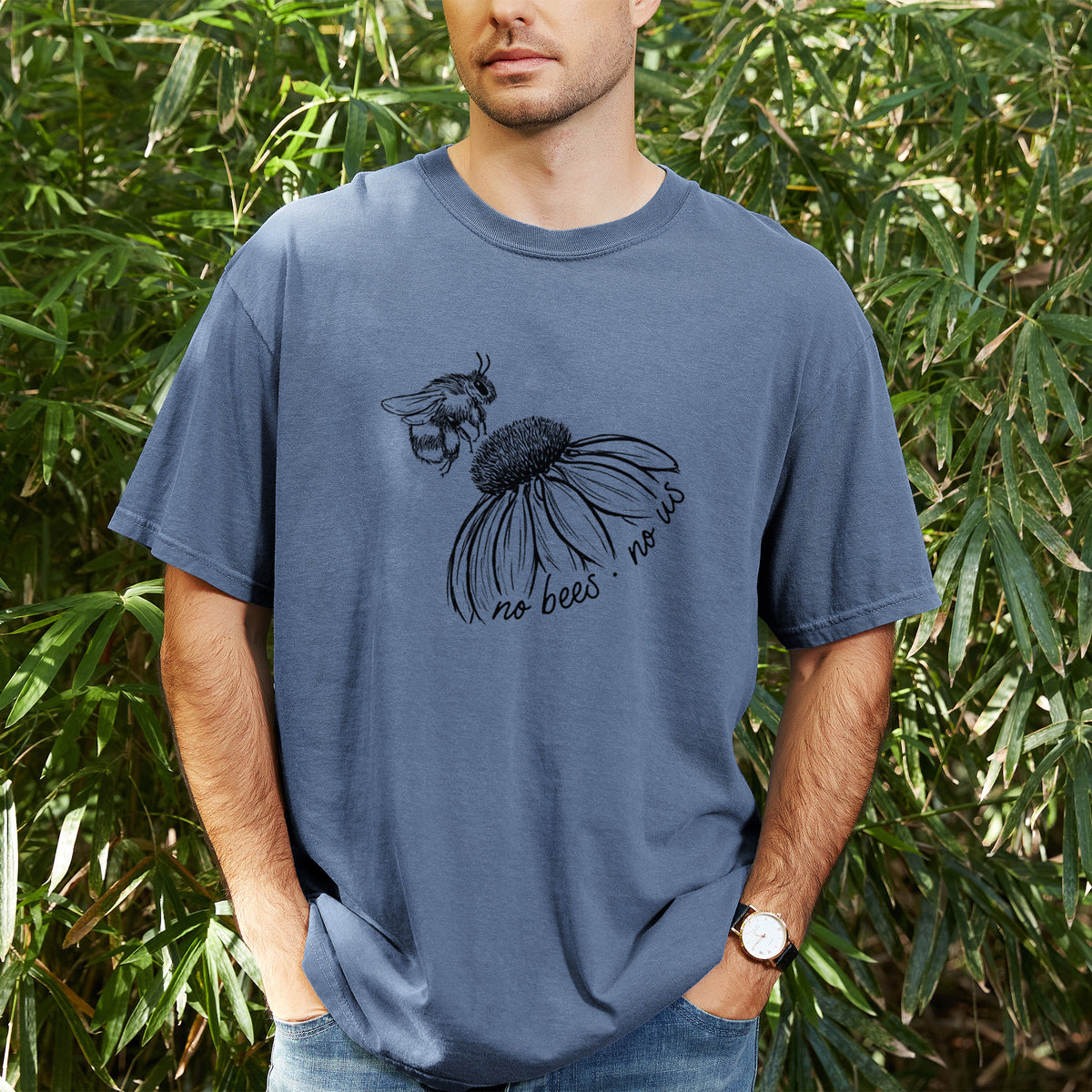 Man wearing a Comfort Colors t-shirt printed with a black ink illustration of a bee flying toward a coneflower, with the handwritten words no bees no us, outdoors.