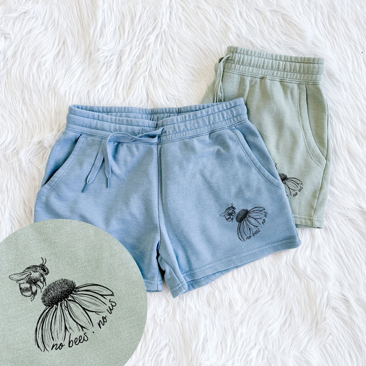 Two pairs of Cali Wave shorts in different colors printed with a black ink illustration of a bee flying toward a coneflower, with the handwritten words no bees no us, with a close-up of the print.