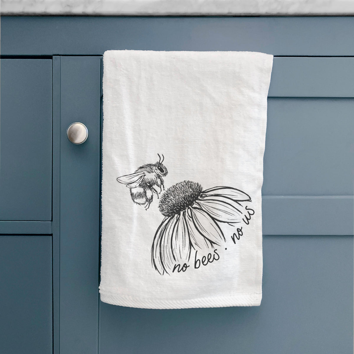 White hand towel printed with a black ink illustration of a bee flying toward a coneflower, with the handwritten words no bees no us, hanging on a cabinet door.