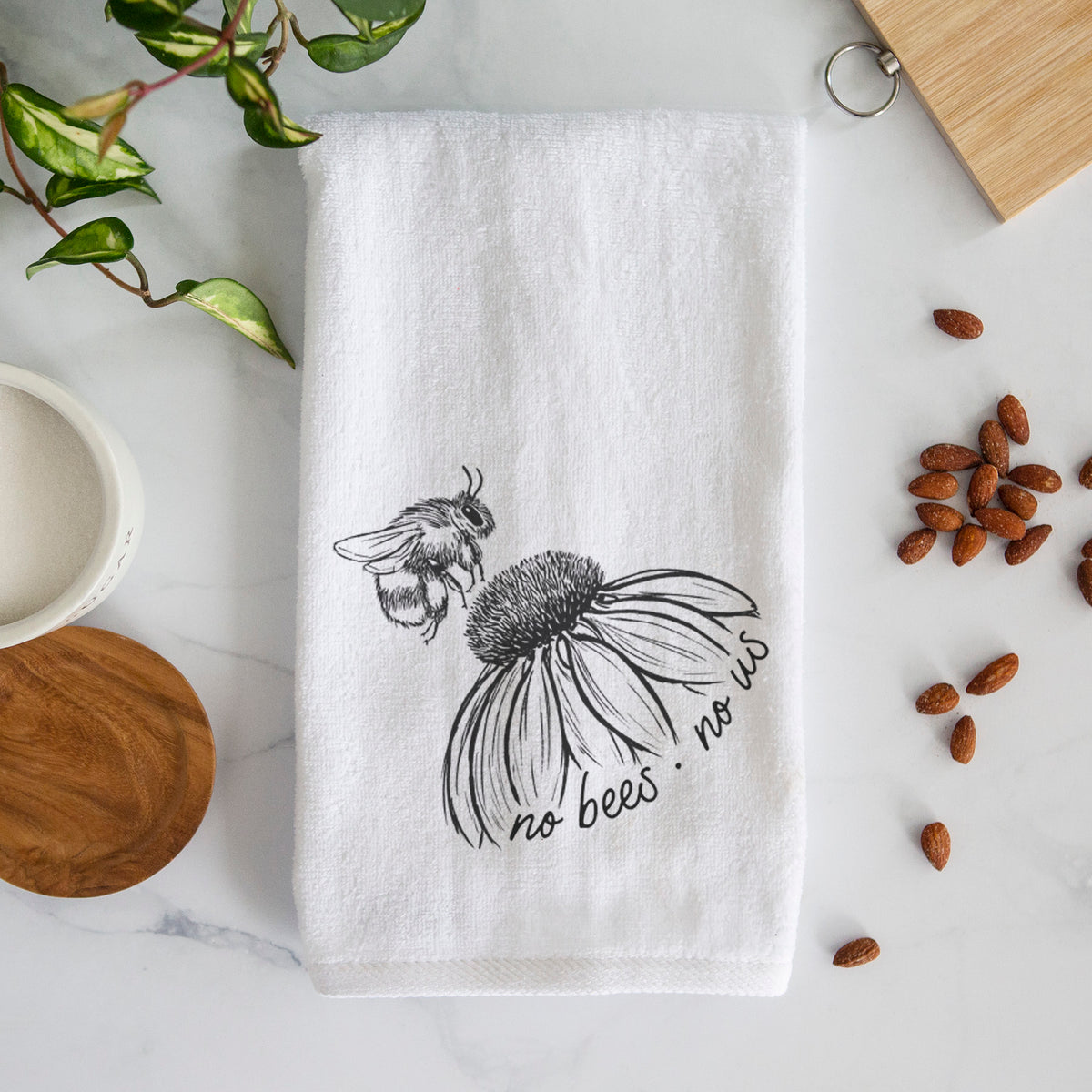 White hand towel printed with a black ink illustration of a bee flying toward a coneflower, with the handwritten words no bees no us.