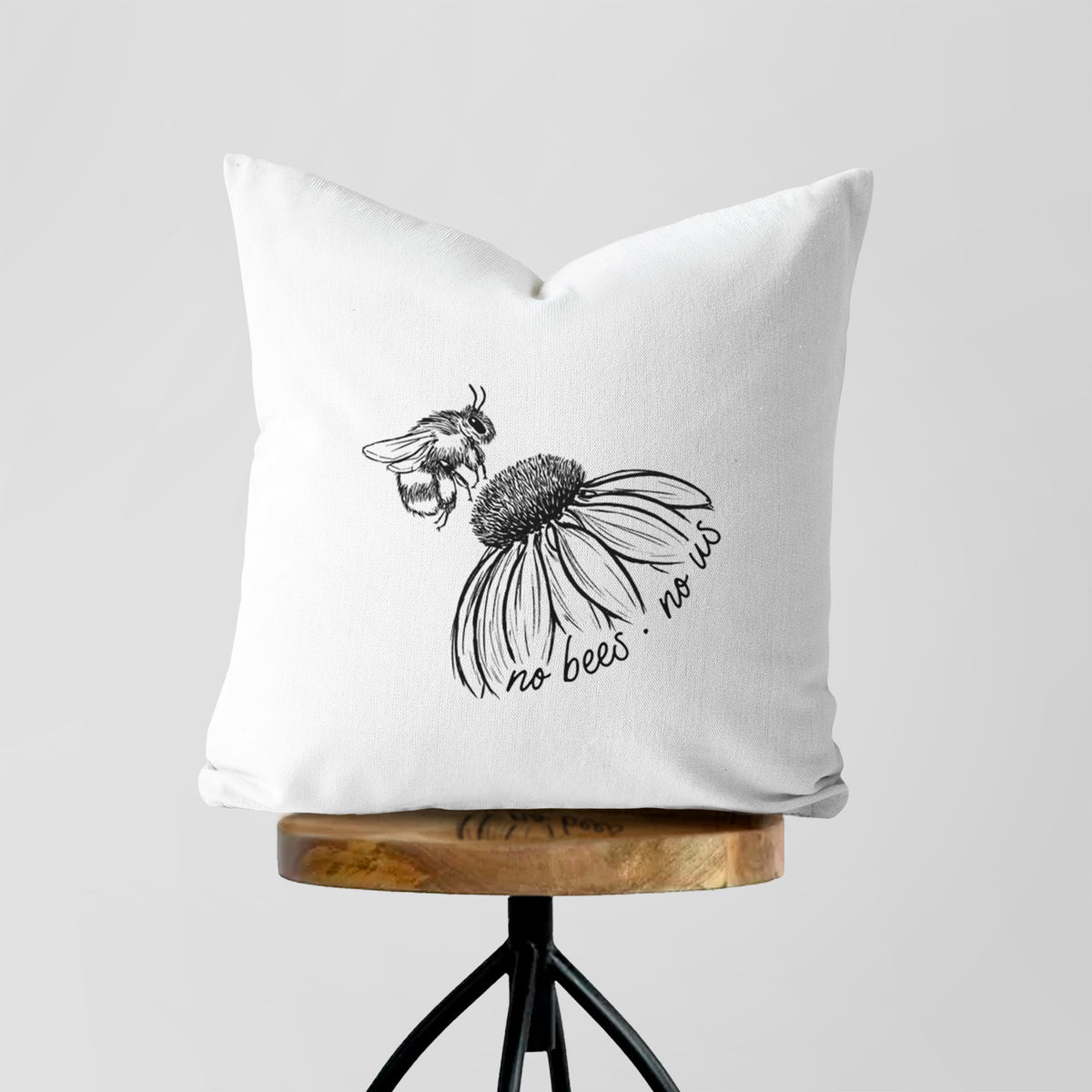 White pillow cover printed with a black ink illustration of a bee flying toward a coneflower, with the handwritten words no bees no us, on a wooden stool.