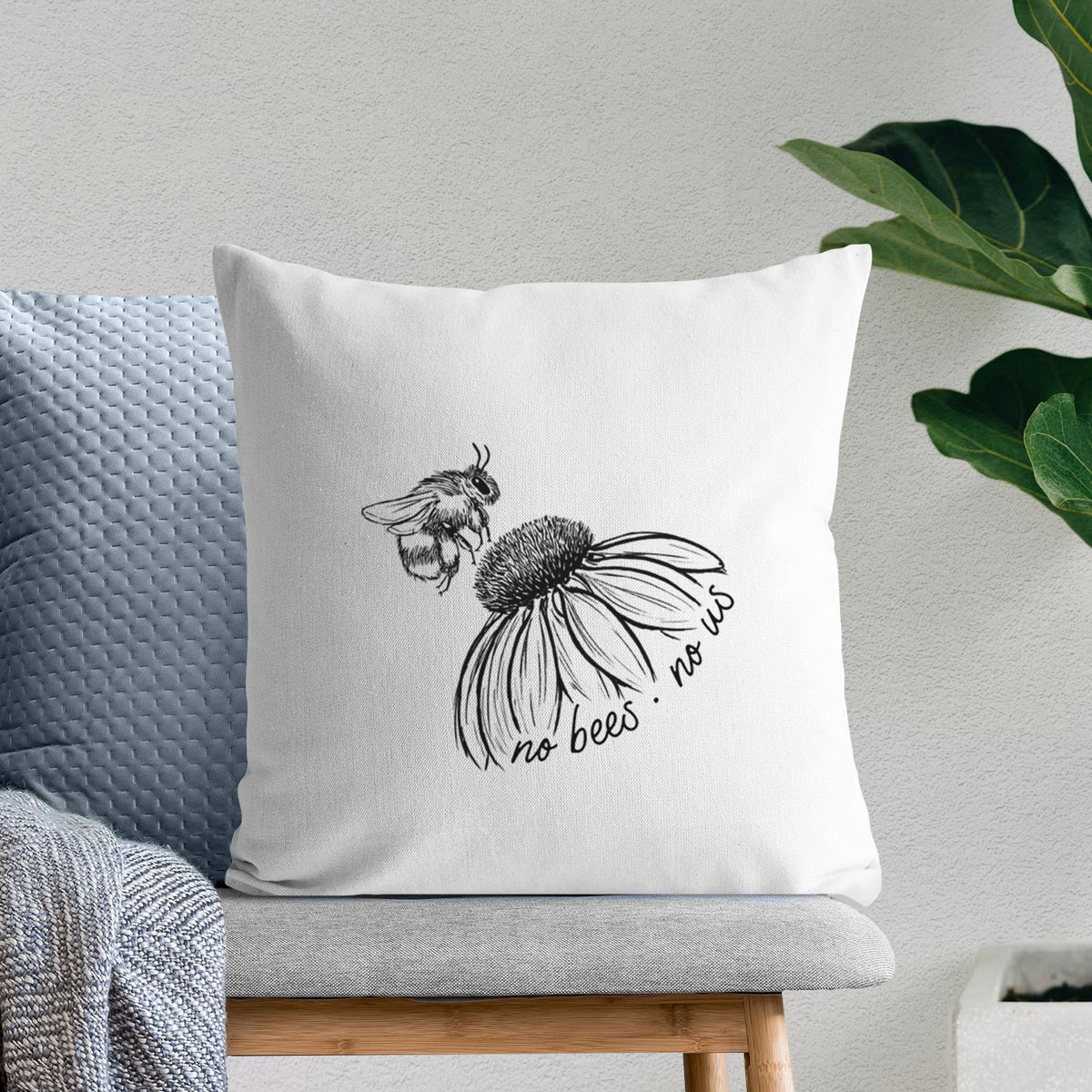 White pillow cover printed with a black ink illustration of a bee flying toward a coneflower, with the handwritten words no bees no us, on a bench beside plants.