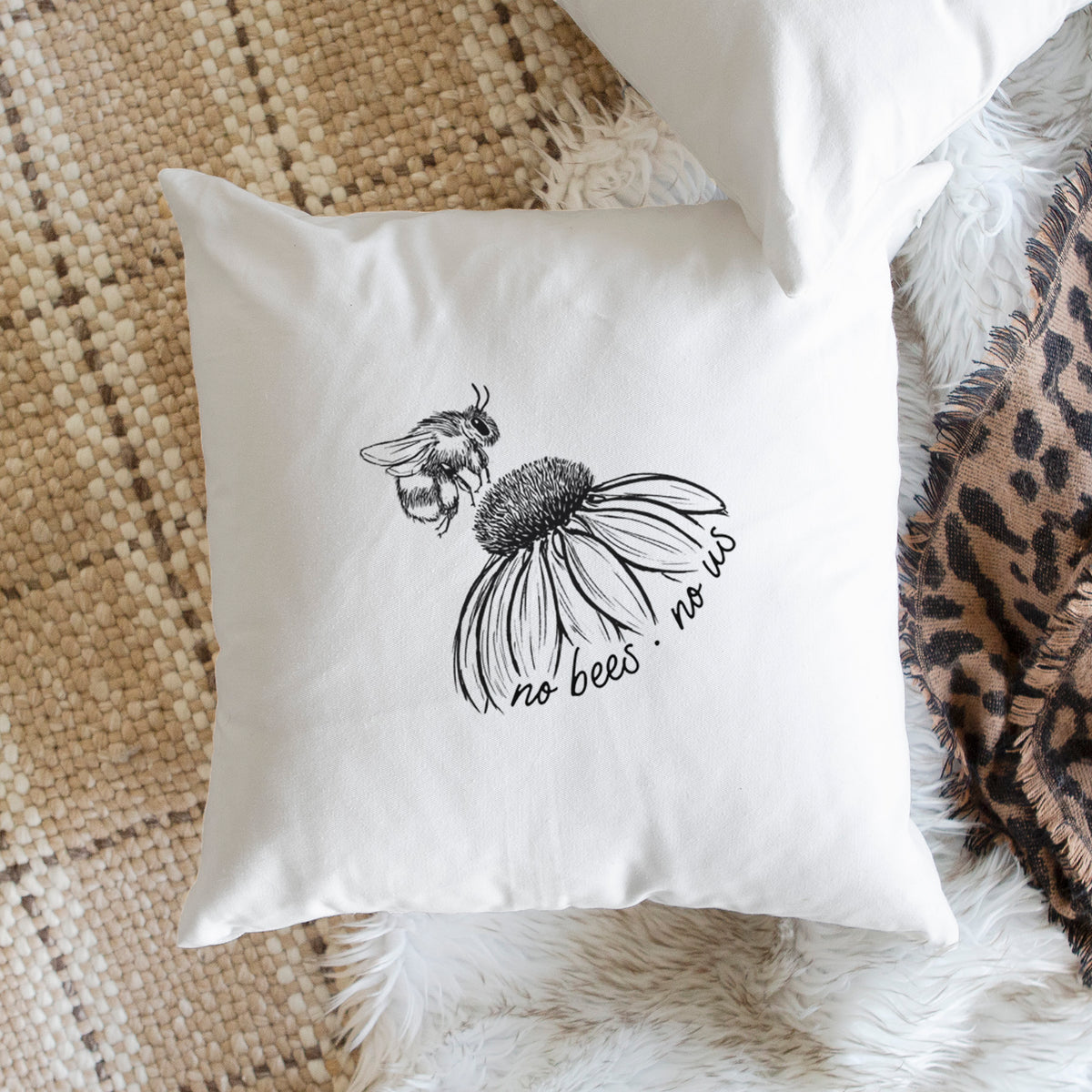 White pillow cover printed with a black ink illustration of a bee flying toward a coneflower, with the handwritten words no bees no us, on a woven chair.