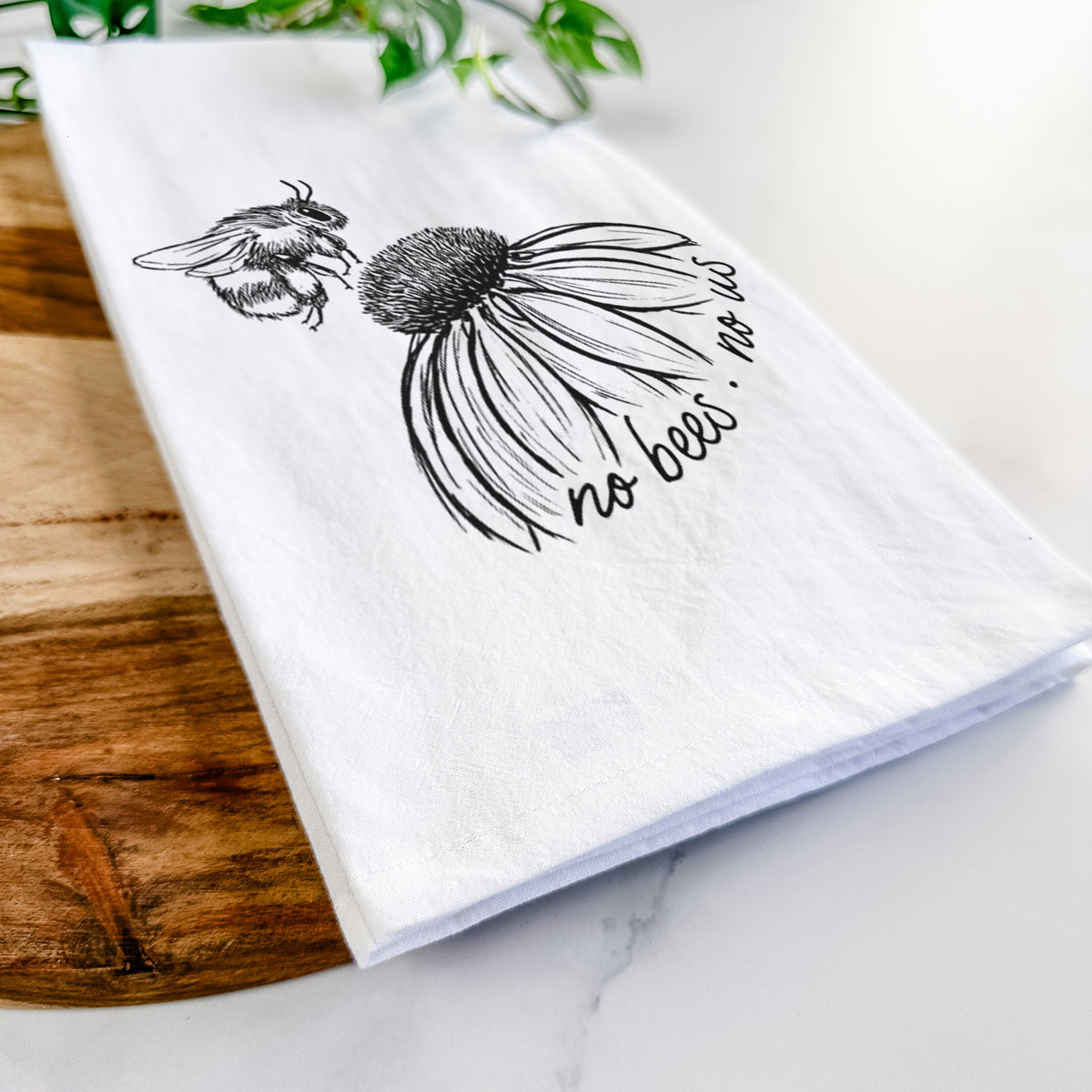Close-up of a folded white tea towel on a wooden board, showing a black ink illustration of a bee flying toward a coneflower, with the handwritten words no bees no us.