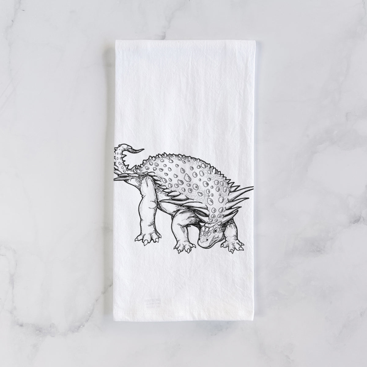 White cotton tea towel laid flat on marble, printed with a black ink illustration of an armored Nodosaurus.