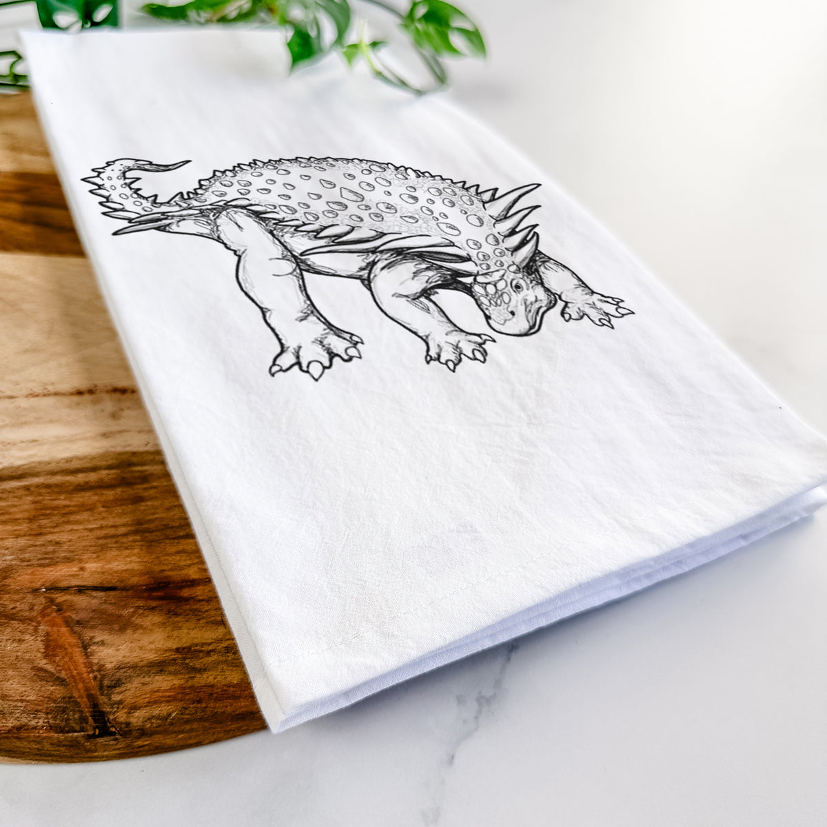 Close-up of a folded white tea towel on a wooden board, showing a black ink illustration of an armored Nodosaurus.