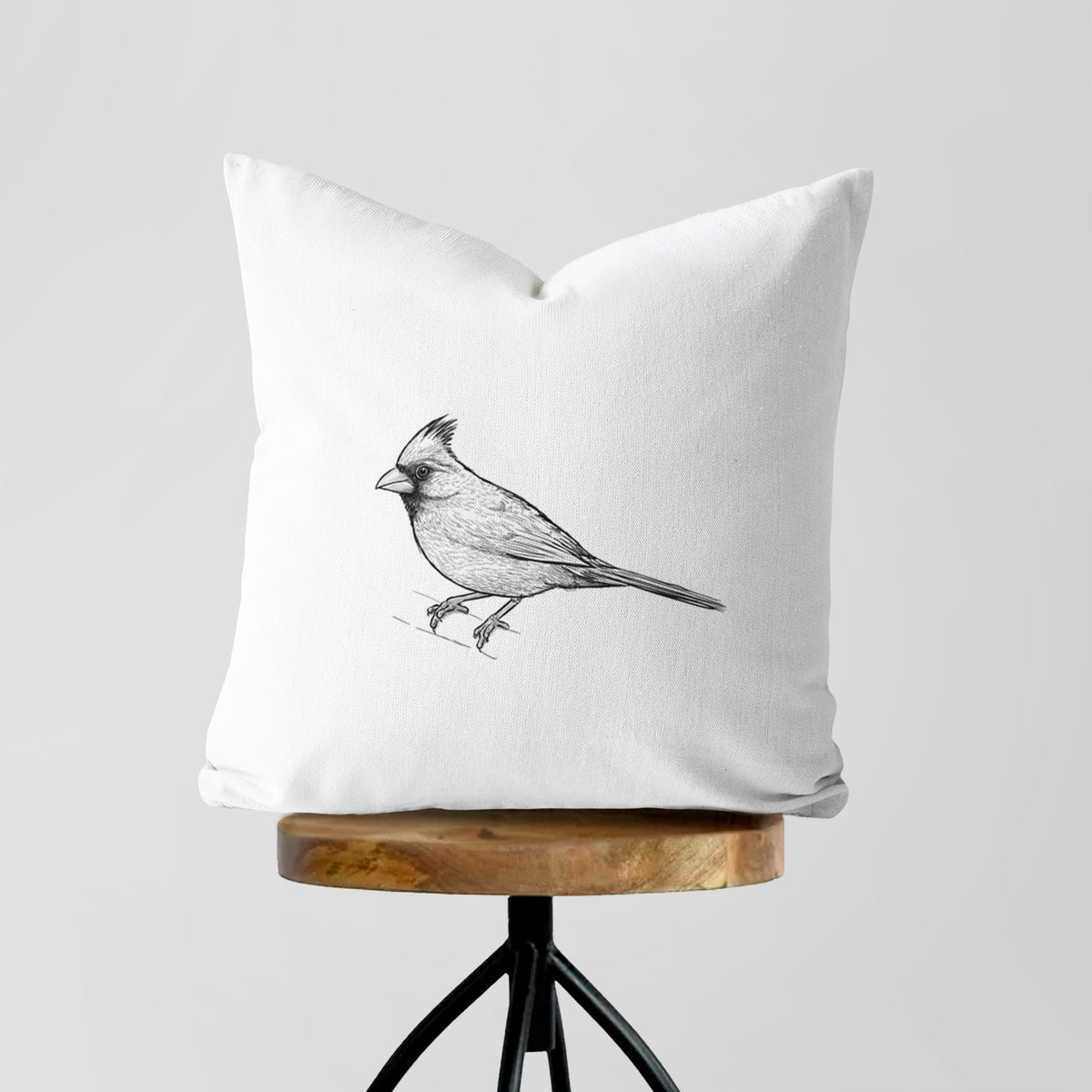 White pillow cover printed with a detailed black ink sketch of a northern cardinal perched with its crest raised, on a wooden stool.
