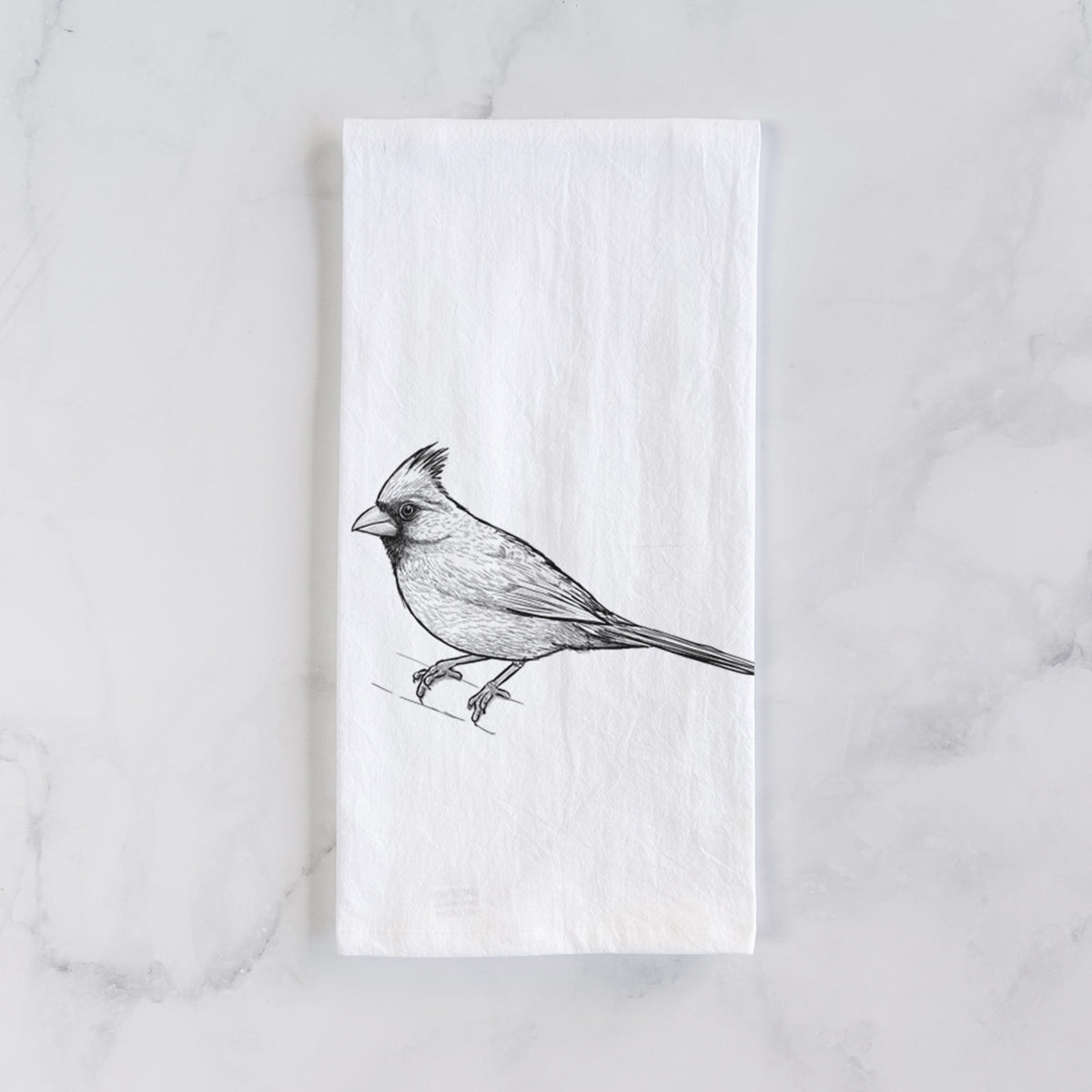 White cotton tea towel laid flat on marble, printed with a detailed black ink sketch of a northern cardinal perched with its crest raised.