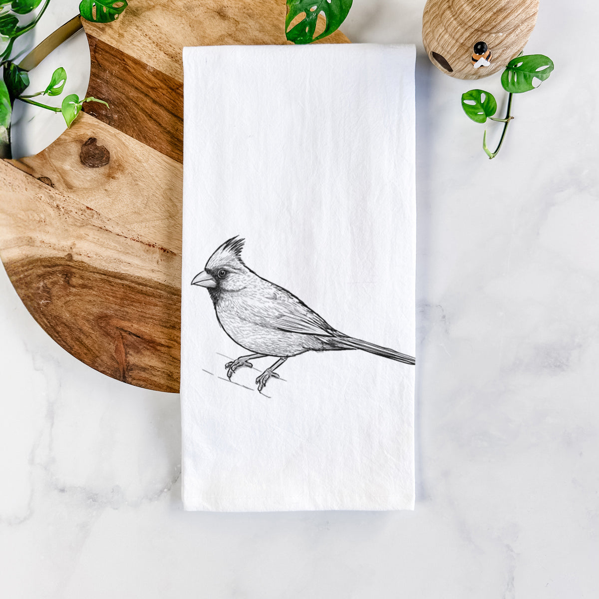 White cotton tea towel printed with a detailed black ink sketch of a northern cardinal perched with its crest raised, on a marble counter beside a round wooden cutting board and greenery.