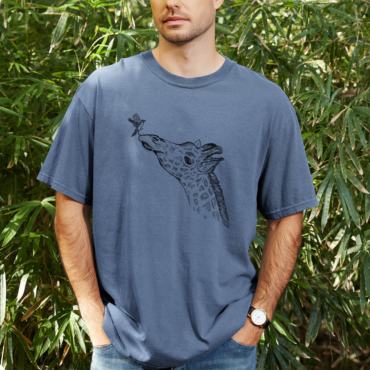 Man wearing a Comfort Colors t-shirt printed with a black ink illustration of a northern giraffe reaching its head up to nibble a flower, outdoors.