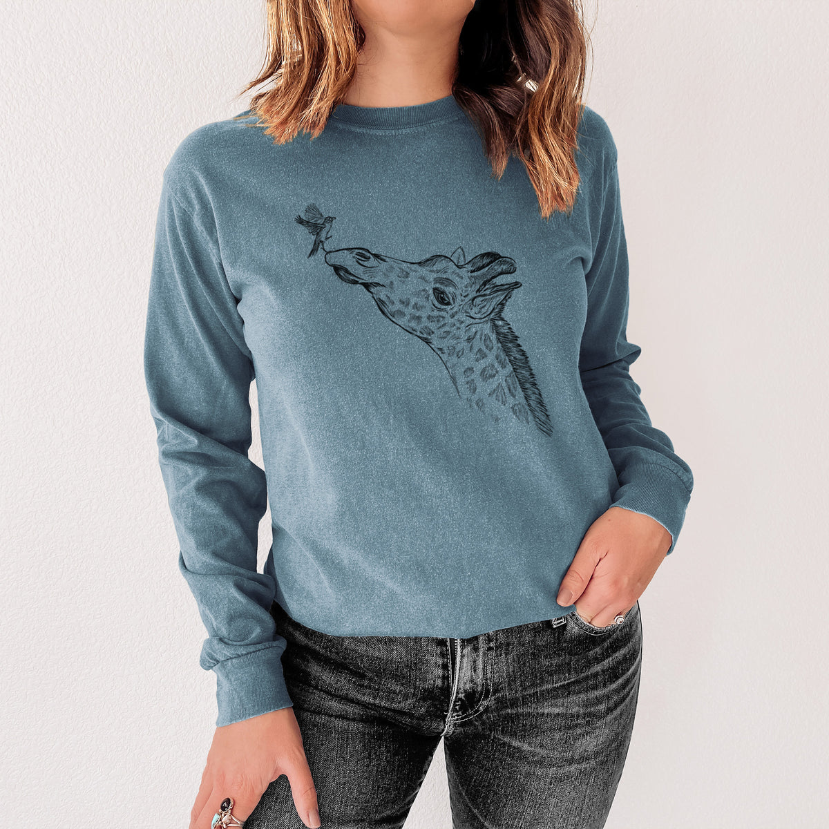 Steel blue Comfort Colors long sleeve shirt printed with a black ink illustration of a northern giraffe reaching its head up to nibble a flower, shown in a lifestyle photo.