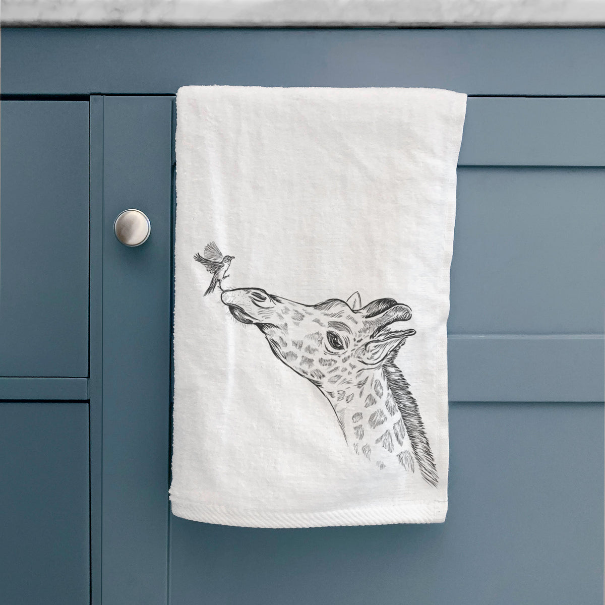 White hand towel printed with a black ink illustration of a northern giraffe reaching its head up to nibble a flower, hanging on a cabinet door.