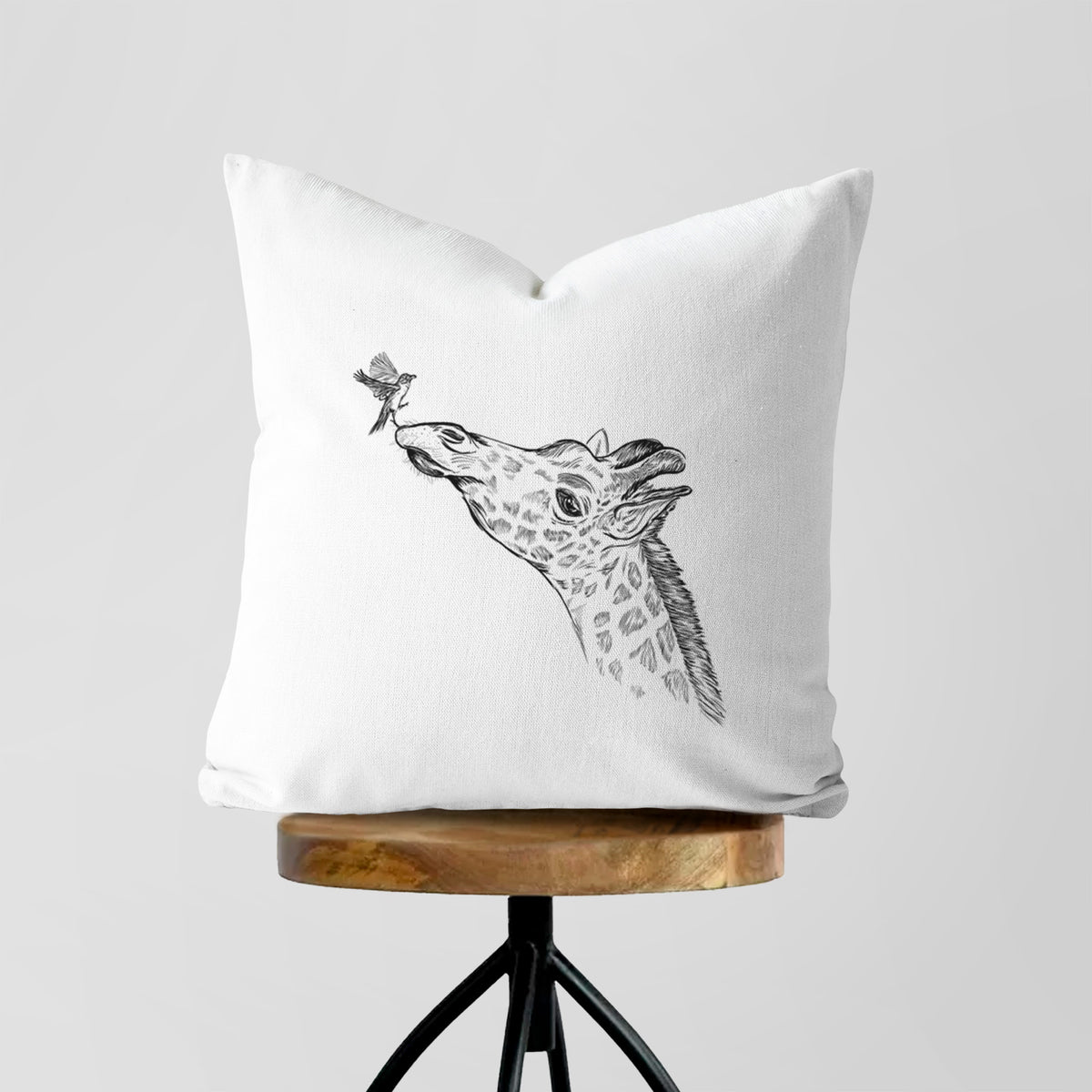 White pillow cover printed with a black ink illustration of a northern giraffe reaching its head up to nibble a flower, on a wooden stool.