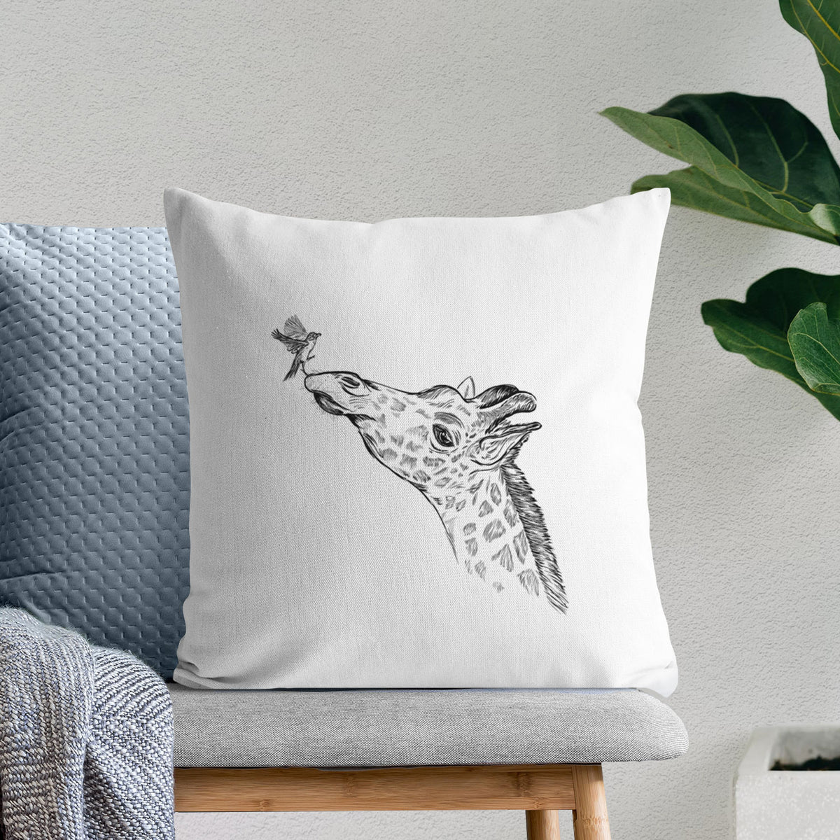White pillow cover printed with a black ink illustration of a northern giraffe reaching its head up to nibble a flower, on a bench beside plants.