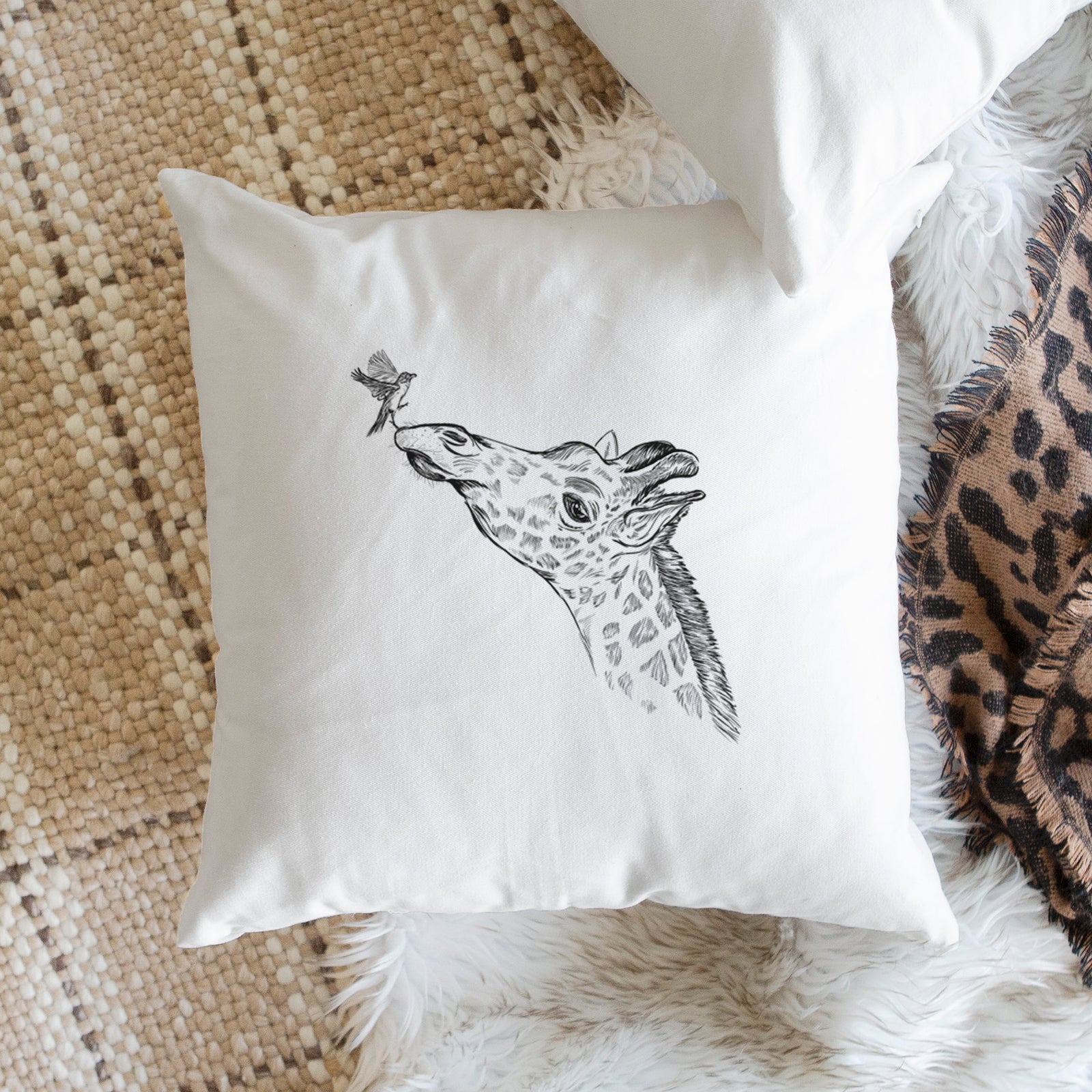 White pillow cover printed with a black ink illustration of a northern giraffe reaching its head up to nibble a flower, on a bench beside plants.