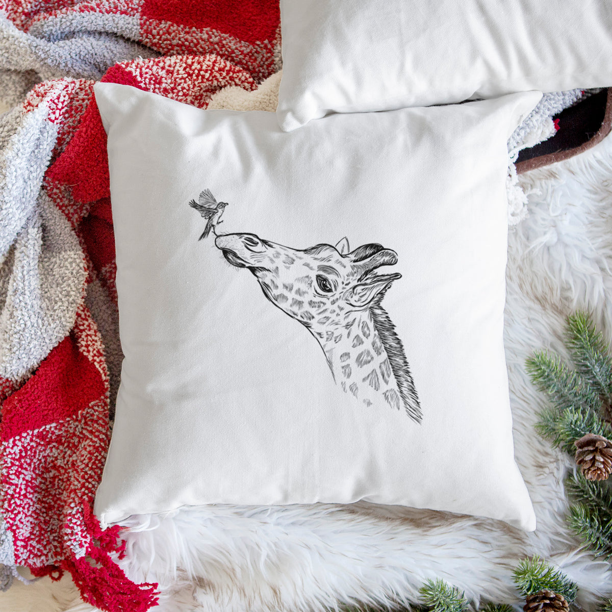 White pillow cover printed with a black ink illustration of a northern giraffe reaching its head up to nibble a flower, on a woven chair.