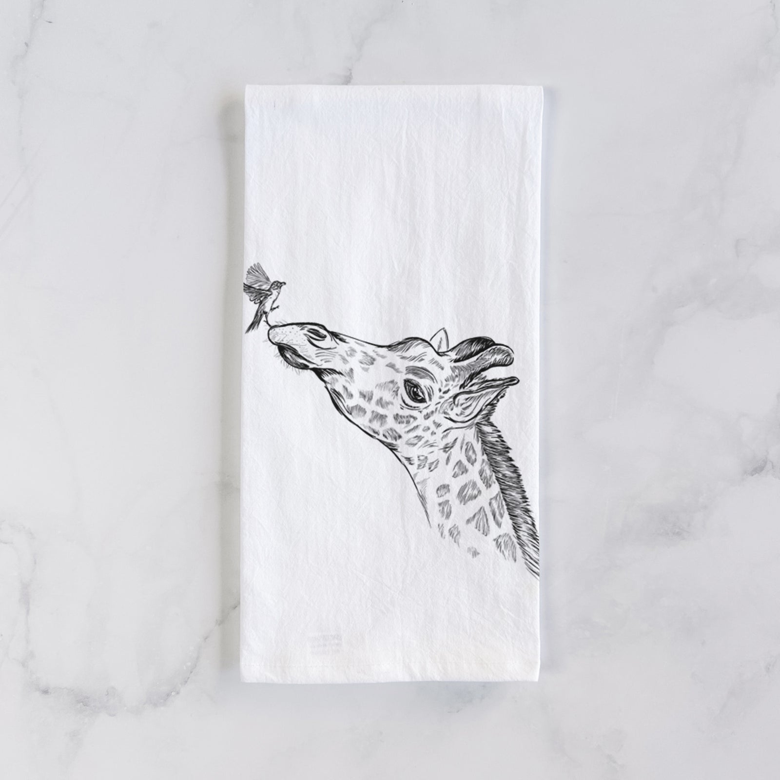 White cotton tea towel printed with a black ink illustration of a northern giraffe reaching its head up to nibble a flower, on a marble counter beside a round wooden cutting board and greenery.