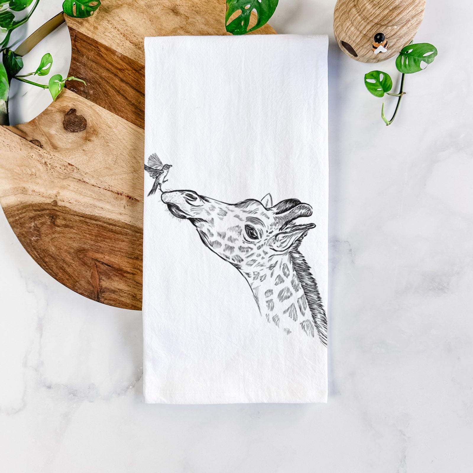 White cotton tea towel printed with a black ink illustration of a northern giraffe reaching its head up to nibble a flower, on a marble counter beside a round wooden cutting board and greenery.