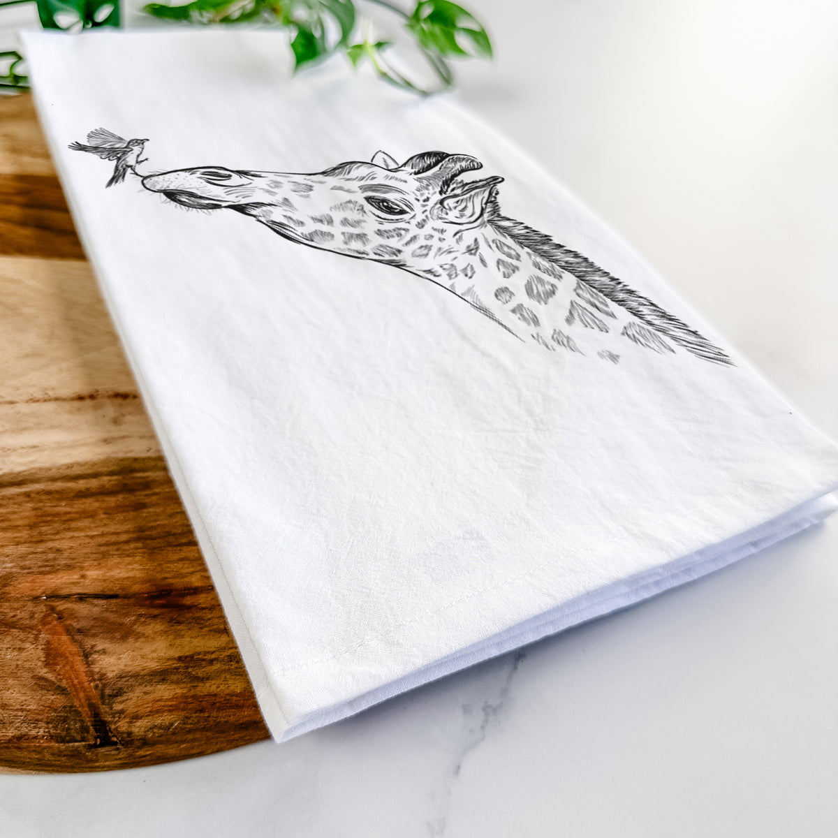 Close-up of a folded white tea towel on a wooden board, showing a black ink illustration of a northern giraffe reaching its head up to nibble a flower.