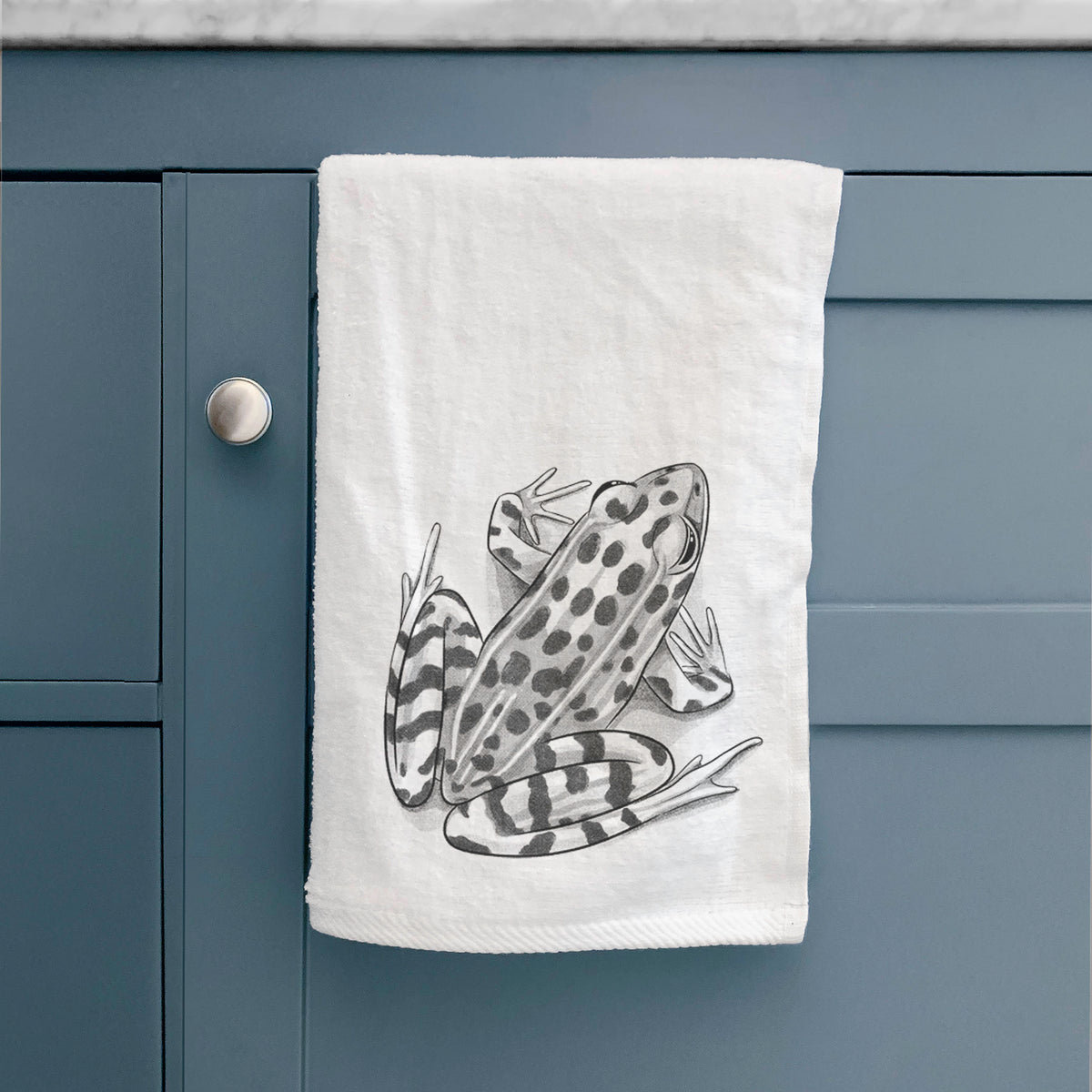 White hand towel printed with a detailed black ink illustration of a spotted northern leopard frog, hanging on a cabinet door.