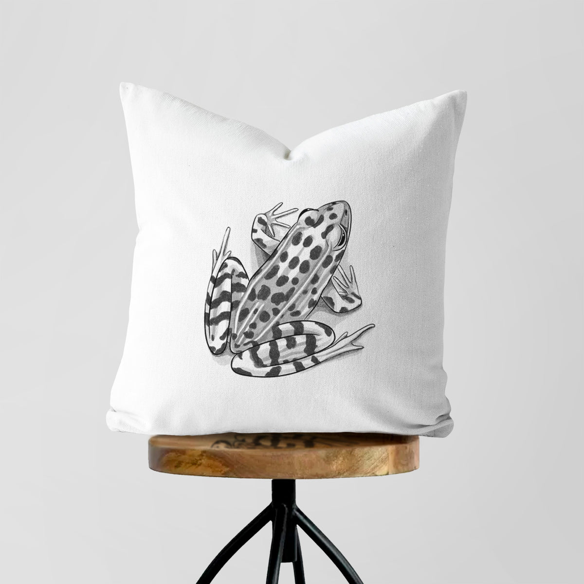 White pillow cover printed with a detailed black ink illustration of a spotted northern leopard frog, on a wooden stool.